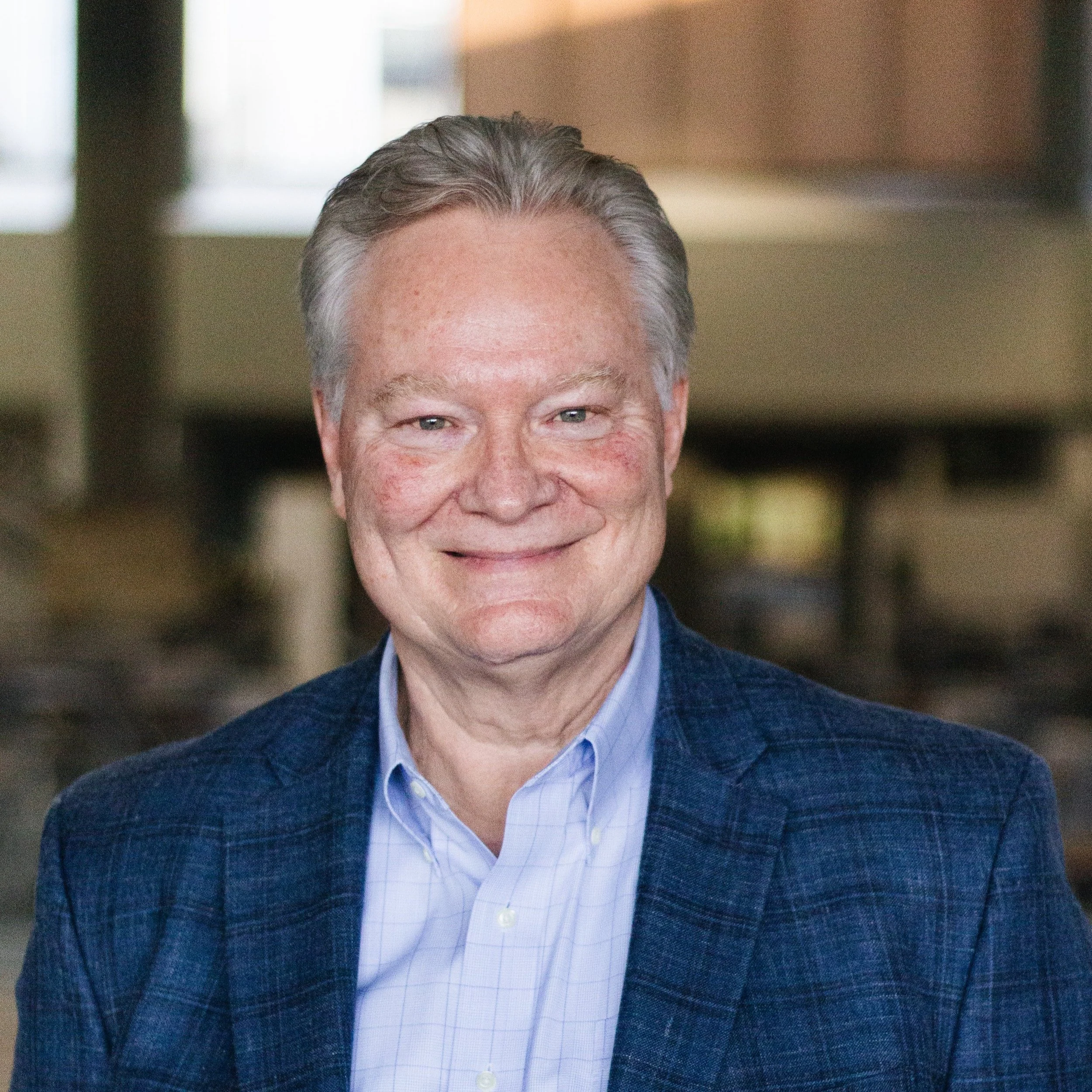 A smiling middle-aged man with gray hair and blue eyes wearing a blue plaid blazer and light blue collared shirt, sitting indoors with a blurred background of windows and furniture.