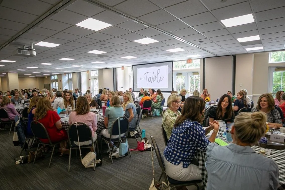 A large gathering of women seated at round tables in a banquet hall, with a screen displaying 'The Table' at the front.