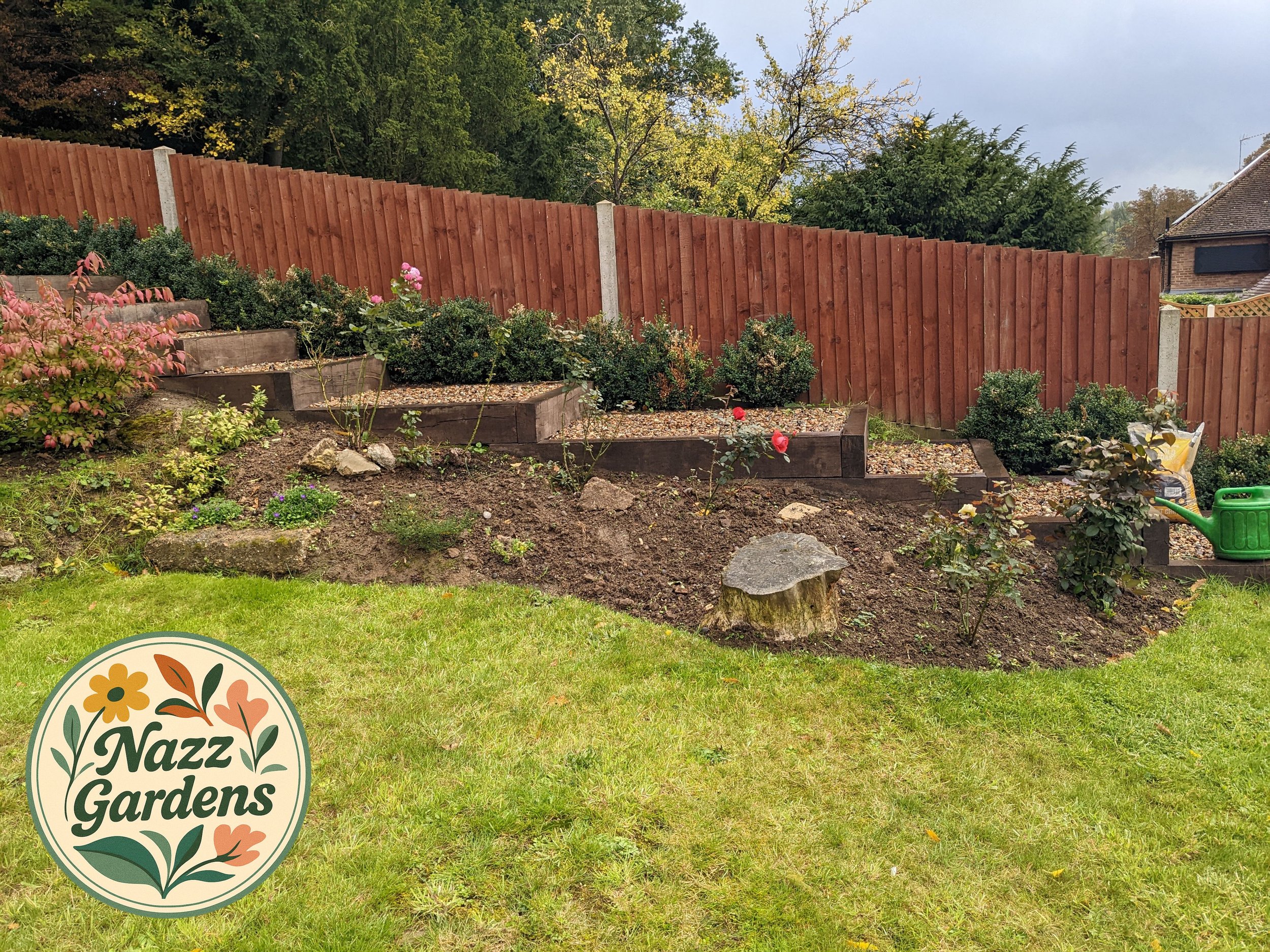 A garden with tiered wooden planters containing flowering bushes and plants, a wooden fence, a grassy lawn in the foreground, and a cloudy sky.
