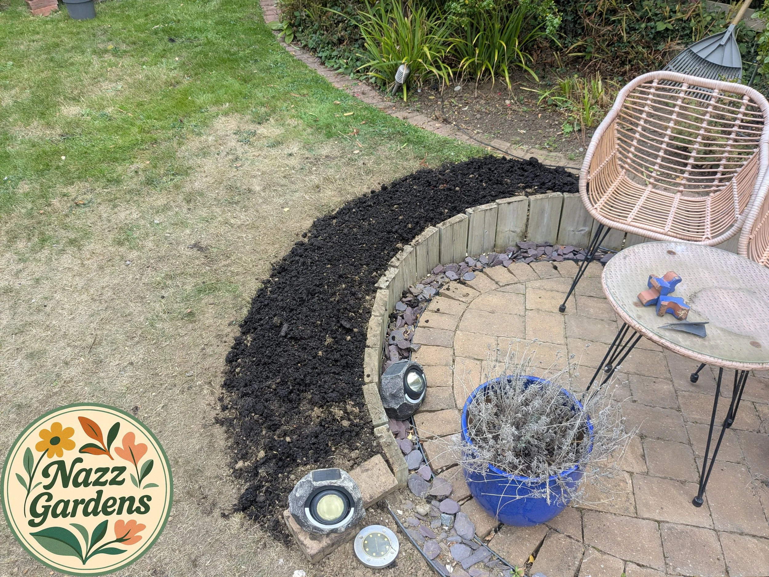 A garden patio with a curved flower bed being prepared with soil, surrounded by wicker chairs and a small glass table. There are garden lights, a blue pot with dried plants, and gardening tools.