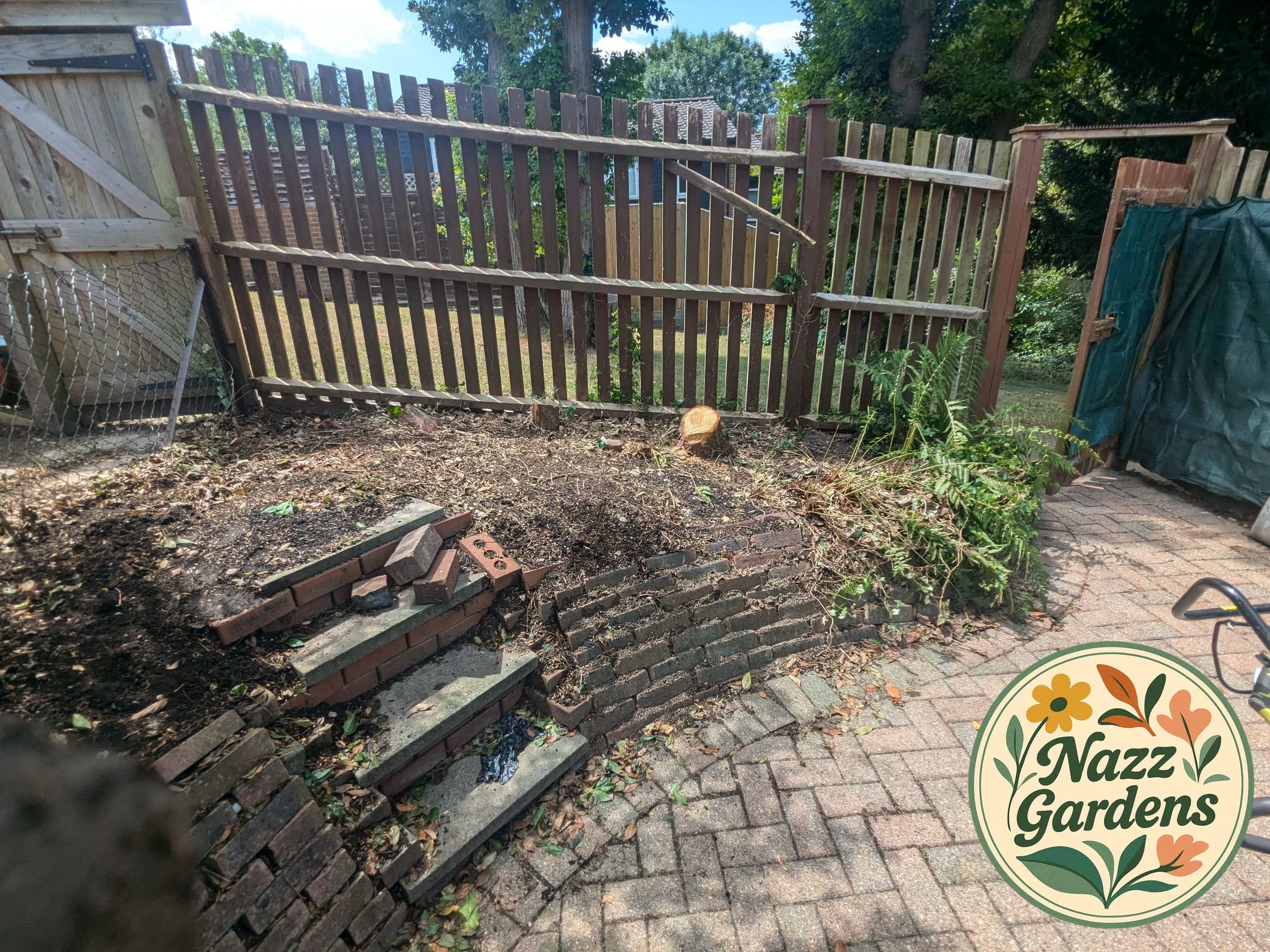 A backyard garden area with a wooden fence, brick steps, and a brick retaining wall. There are some plants and soil, with gardening tools and equipment nearby.
