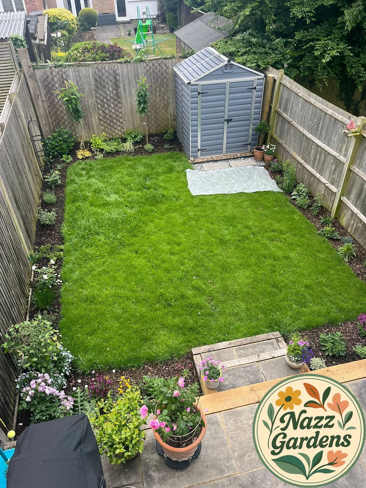 A backyard garden with a green lawn, flower beds along three sides, a gray shed, potted plants, and a paved patio area, viewed from above.