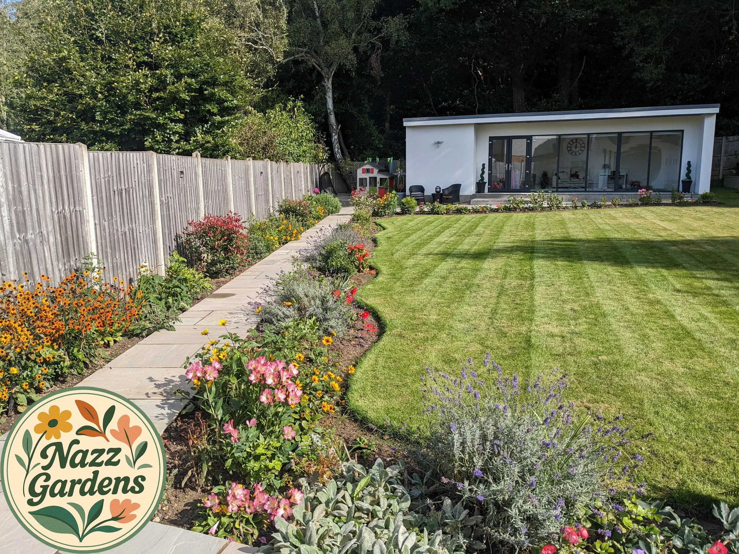 A modern backyard with a well-maintained lawn, colorful flower beds along a curved stone pathway, a wooden fence, and a white house with large glass windows and a patio area with outdoor furniture.