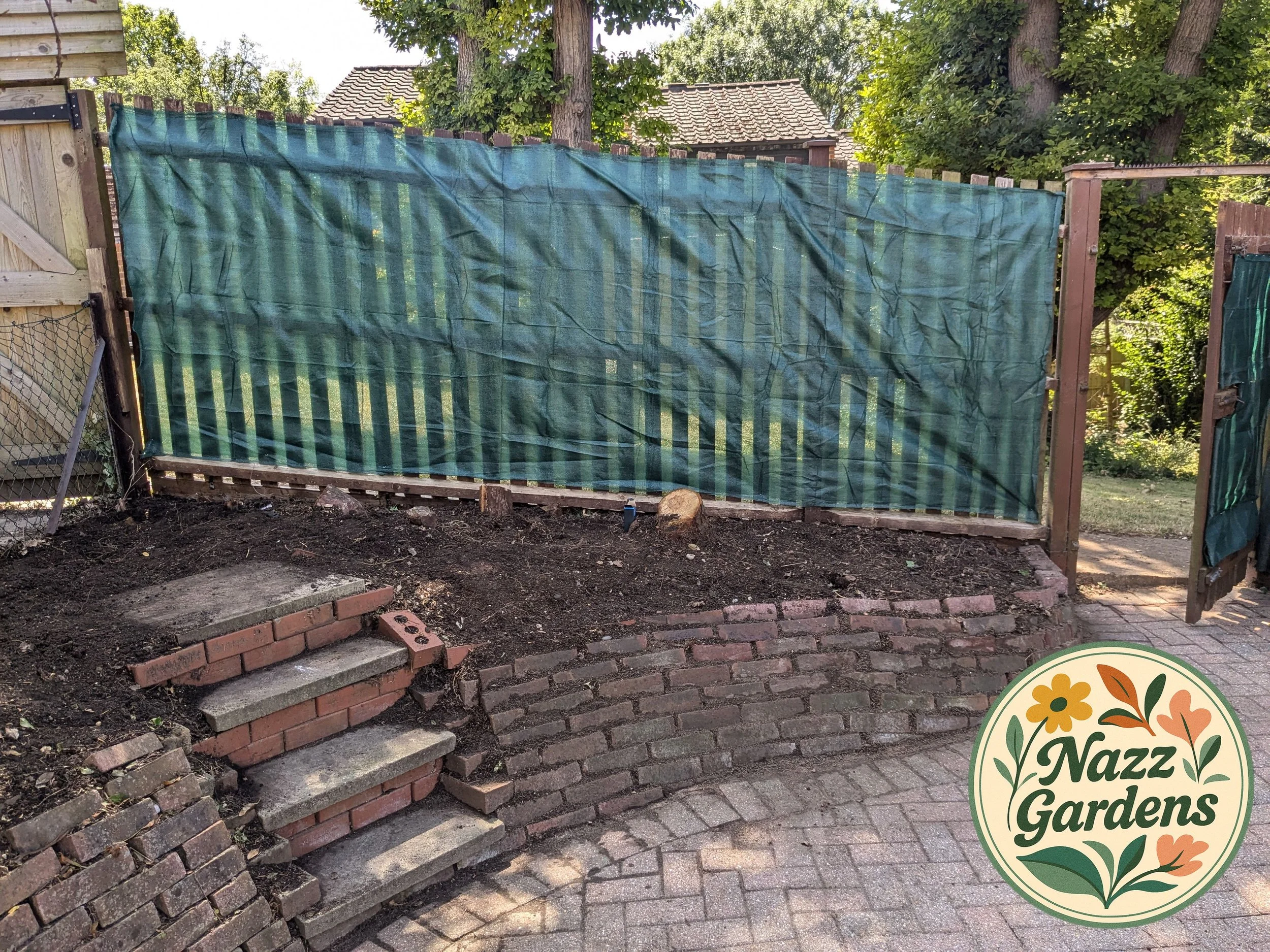 A backyard garden area with a dirt bed surrounded by a brick retaining wall and a wooden fence with a green tarp covering part of it, and a small staircase made of bricks and concrete steps leading up to the garden bed. There are trees and houses in the background.