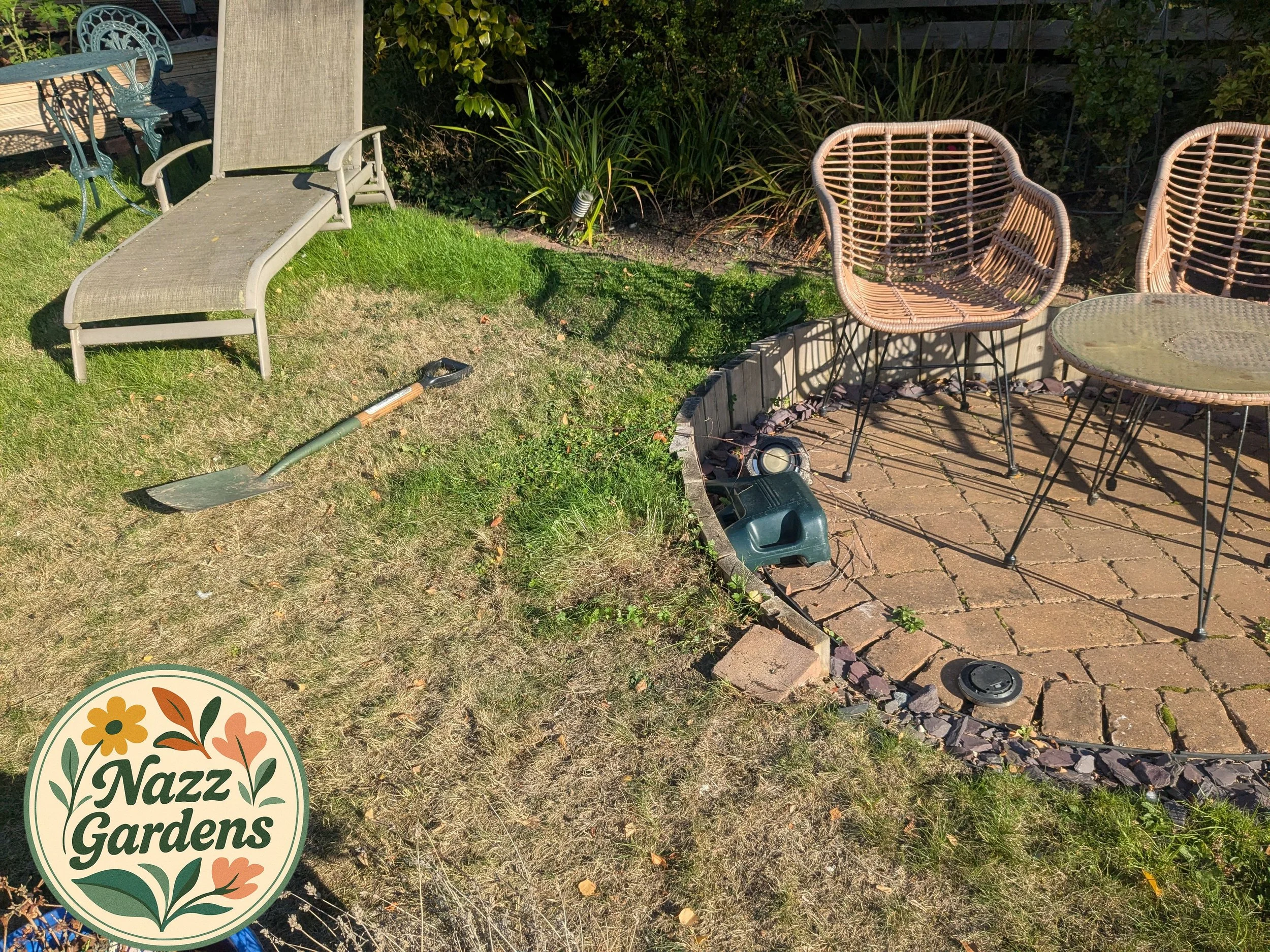 Backyard garden with patio furniture, including a beige lounge chair, rattan chairs, and a glass-top table, with a garden bed and a mix of green and dry grass, and a shovel lying on the grass. There is also a garden watering can and a black lid on the brick patio. The image has a Nazz Gardens logo in the bottom left corner.