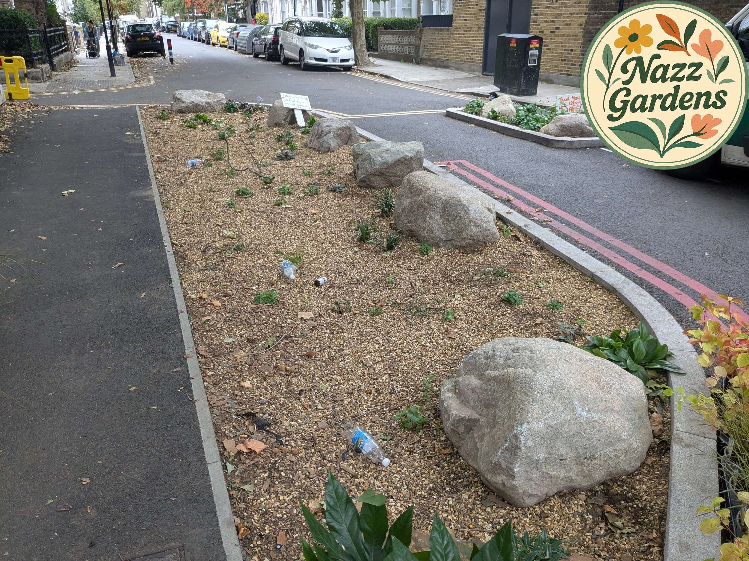 A small urban garden bed with large rocks, sparse plants, and some litter, situated on a street with parked cars and a sidewalk, with a sign reading 'Nazz Gardens' in the top right corner.