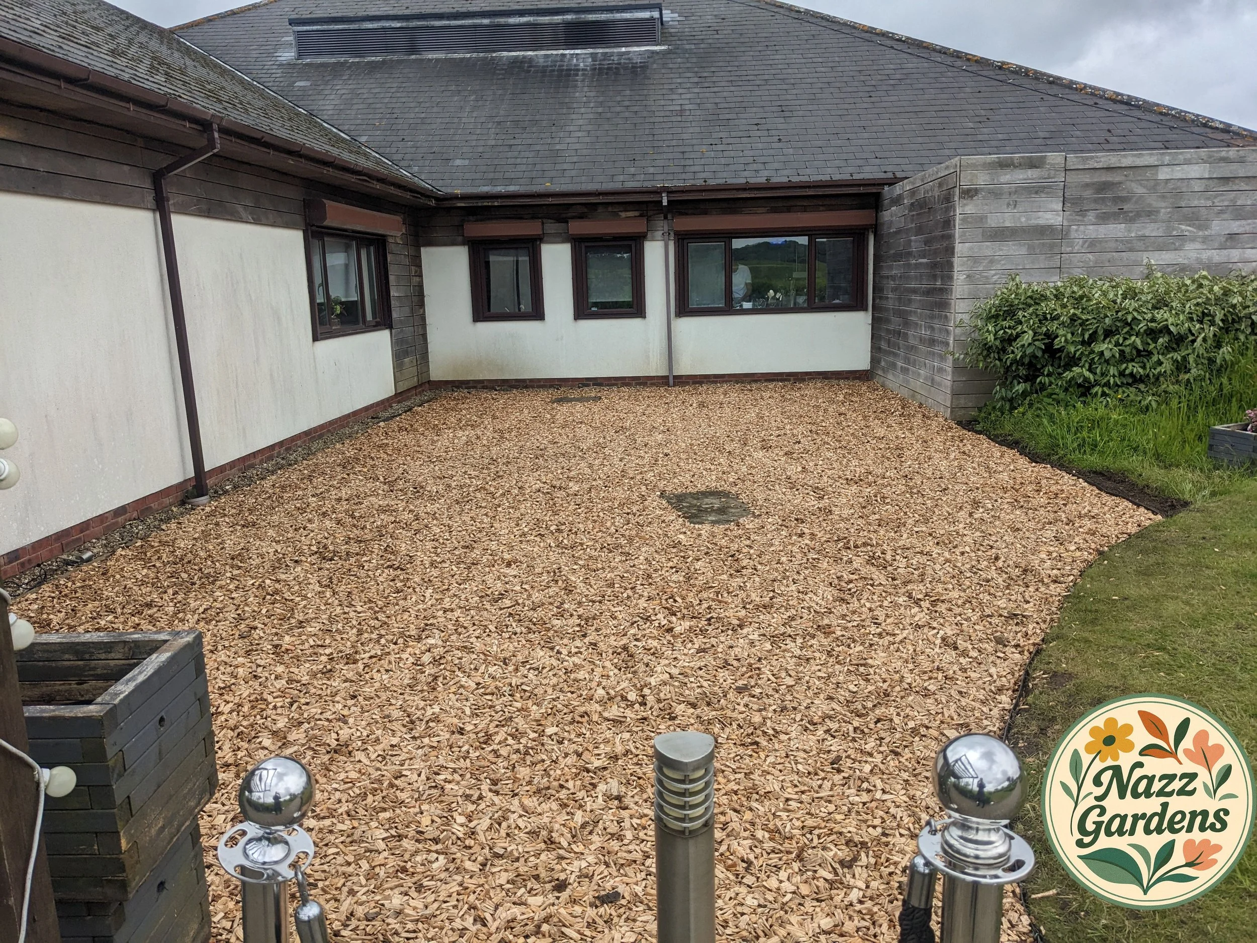 Backyard with a gravel-covered area, house with white walls, brown window frames, and wooden panelling, and a garden area with green plants, a lawn.