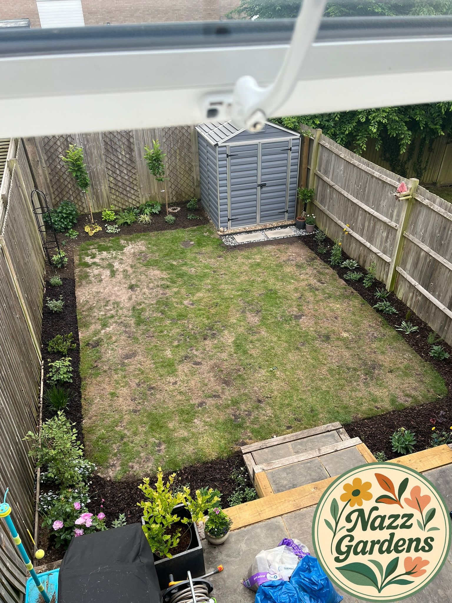 A backyard with a small lawn, garden beds along the fence, a gray garden shed, and a patio area in the foreground with potted plants and gardening tools.