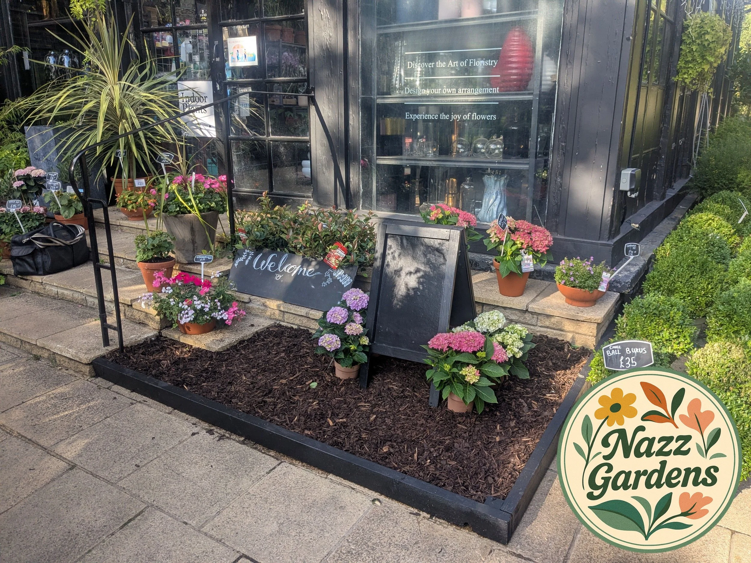 Flower display outside with potted plants and flowers, black chalkboard signs, and a black building with decorative items inside.