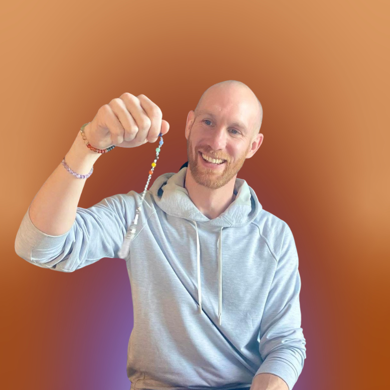A smiling man wearing a light gray hoodie holds up a colorful beaded bracelet against a warm, blurred background.