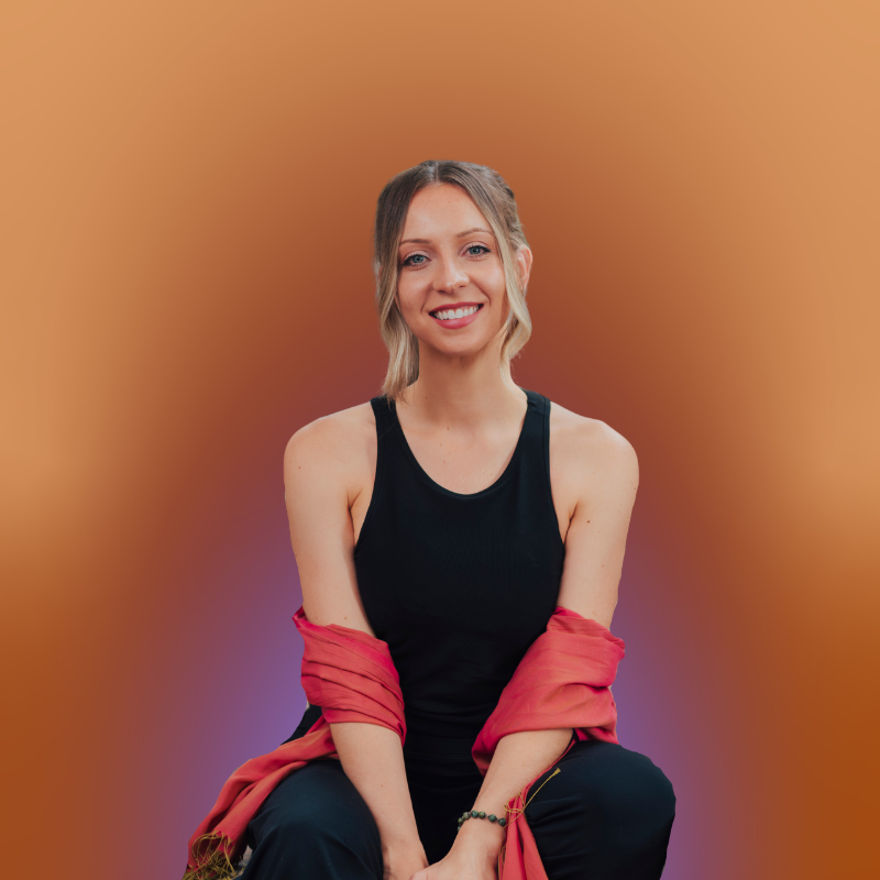 A young woman with blonde hair styled in loose waves, wearing a black sleeveless top and black pants, sitting against a gradient orange background with a red jacket draped over her arms and a bracelet on her left wrist, smiling at the camera.