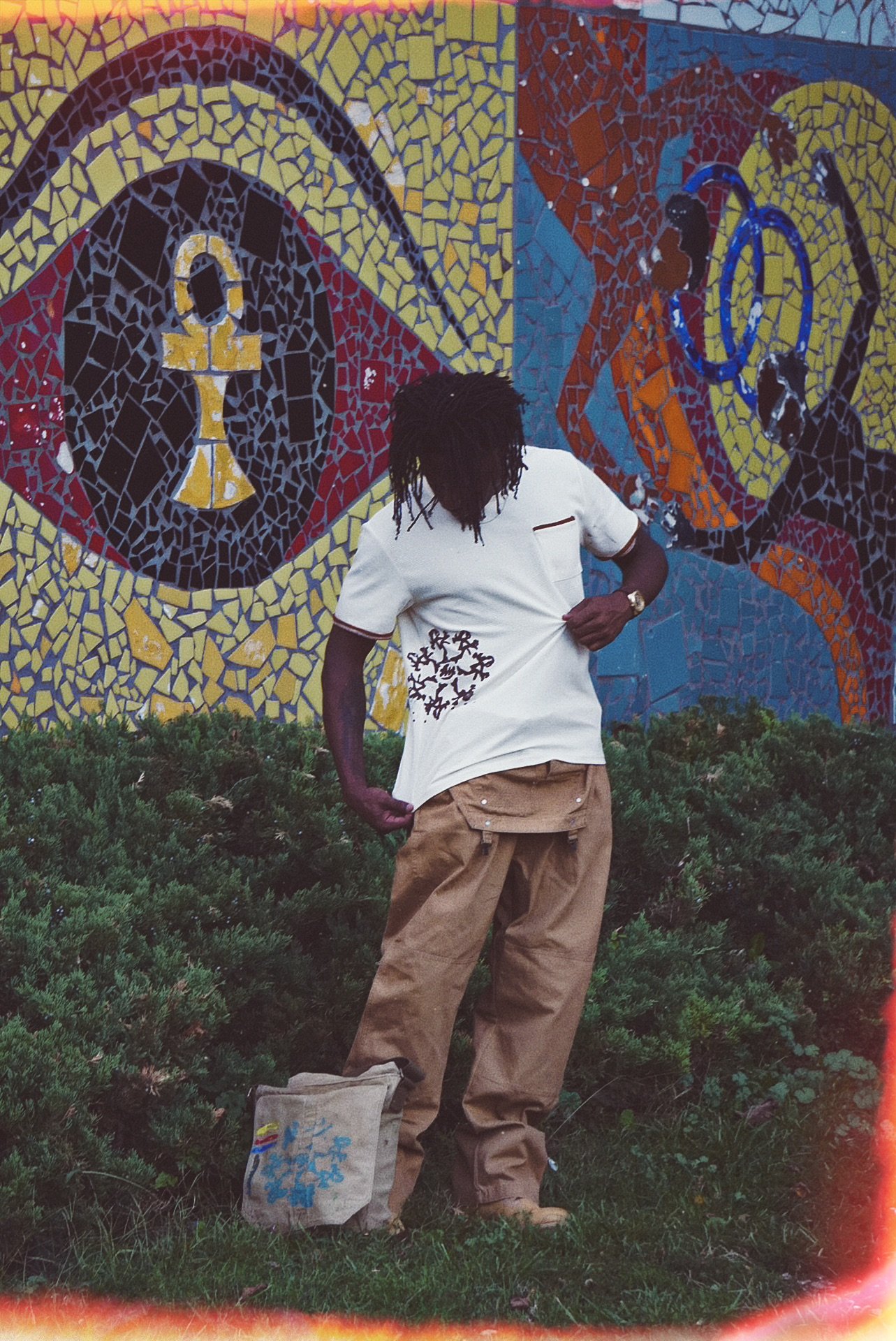 A person with dreadlocks standing outdoors in front of a colorful mosaic mural displaying two stylized faces and symbols. The person is wearing a white t-shirt with a pattern, tan pants, and a watch, and has a beige bag on the ground nearby.