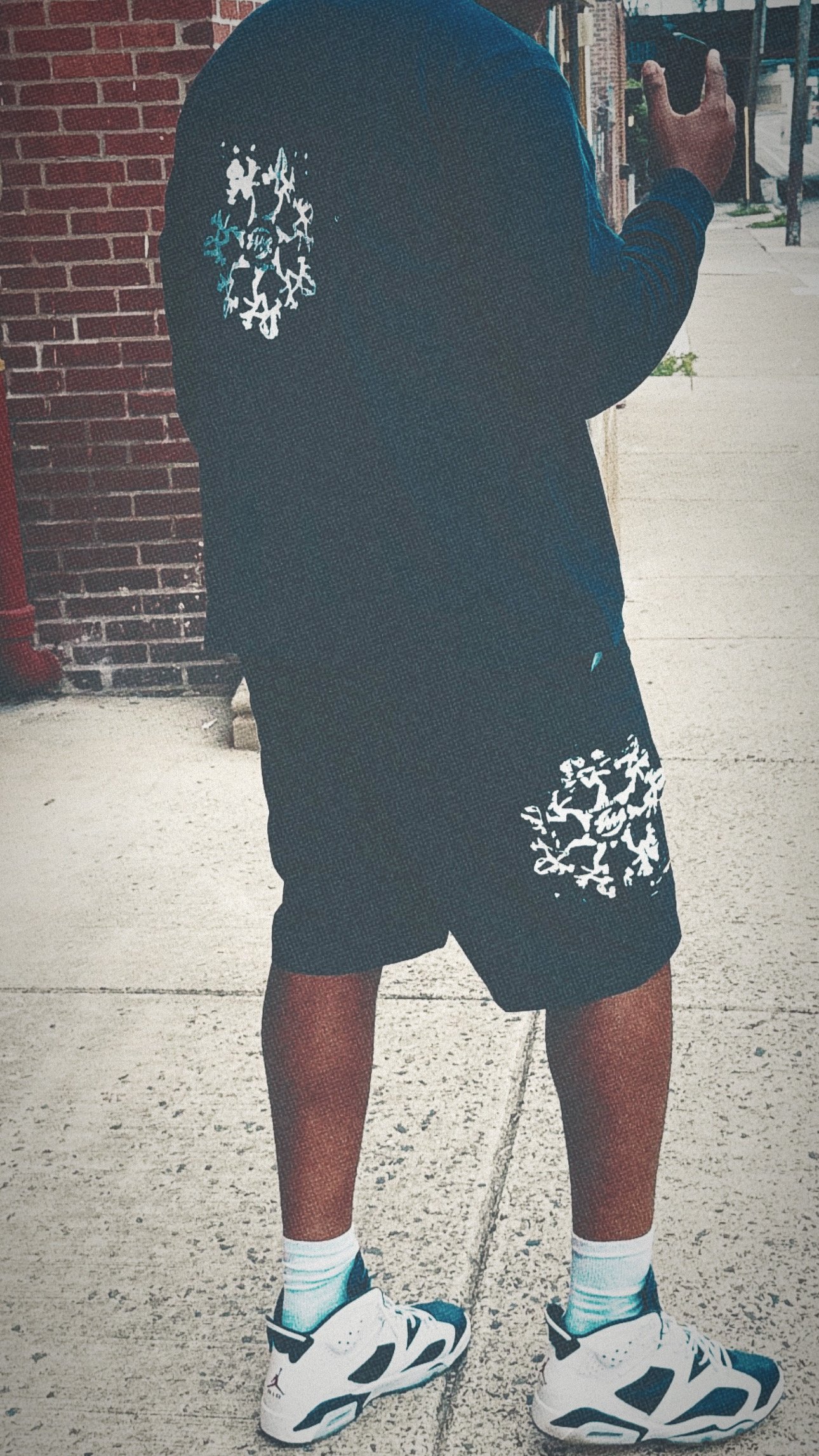 Part of a person's body, showing legs in shorts, a dark sweatshirt with white and blue patterns, white sneakers, and ankle socks, standing on a sidewalk next to a brick wall.