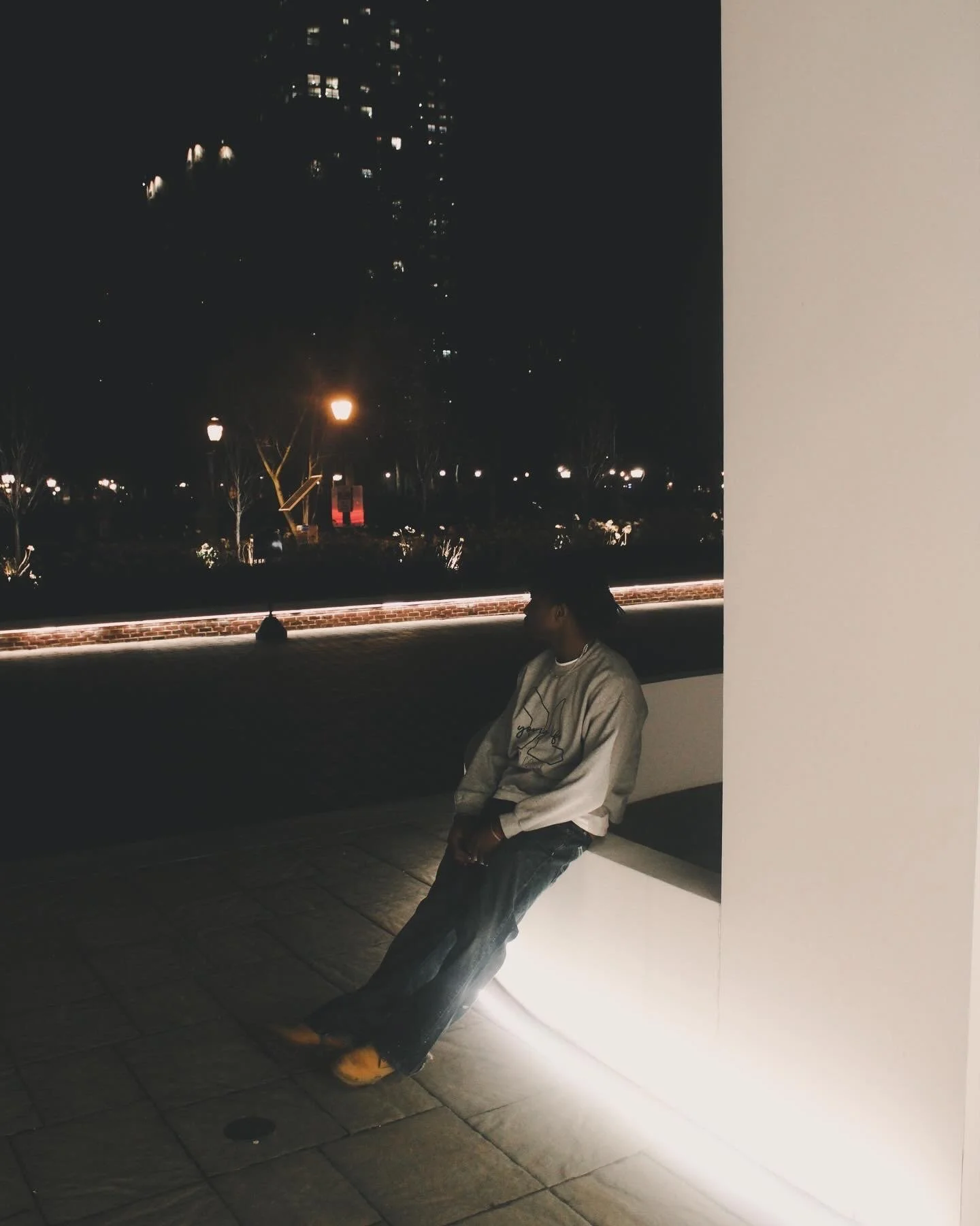 Person sitting on a ledge outside at night, with city lights and buildings in the background, wearing a beige hoodie and jeans.