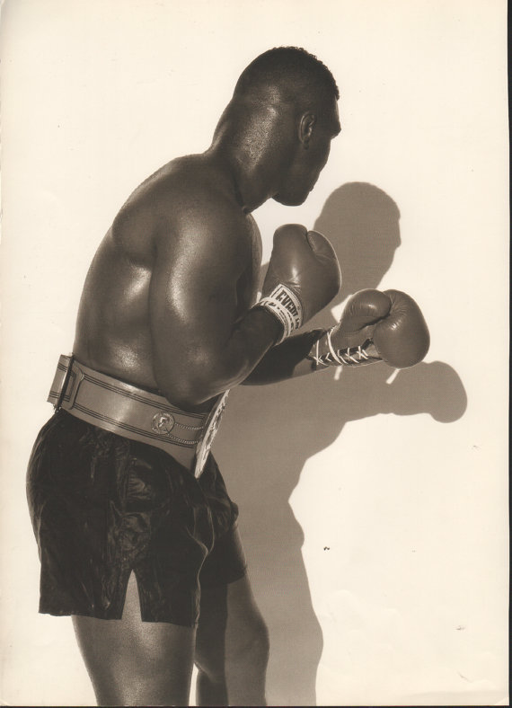 Black and white photograph of a boxer in a fighting stance, wearing boxing gloves, boxing trunks, and a championship belt, against a plain background.