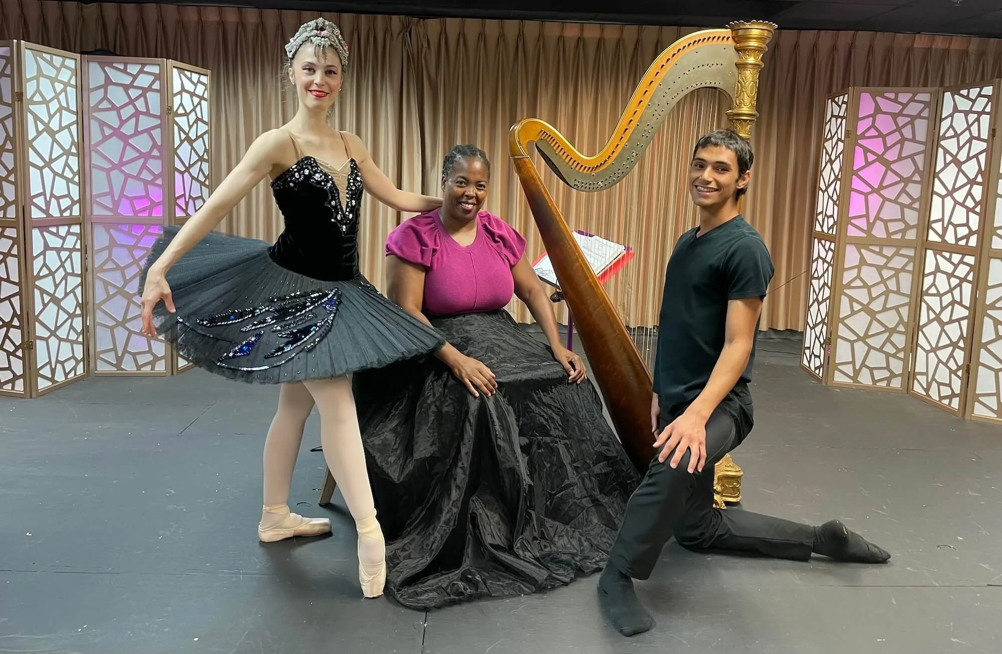Ballet dancer in a black sequined tutu and crown posing on stage with a harpist and a male dancer alongside a concert harp