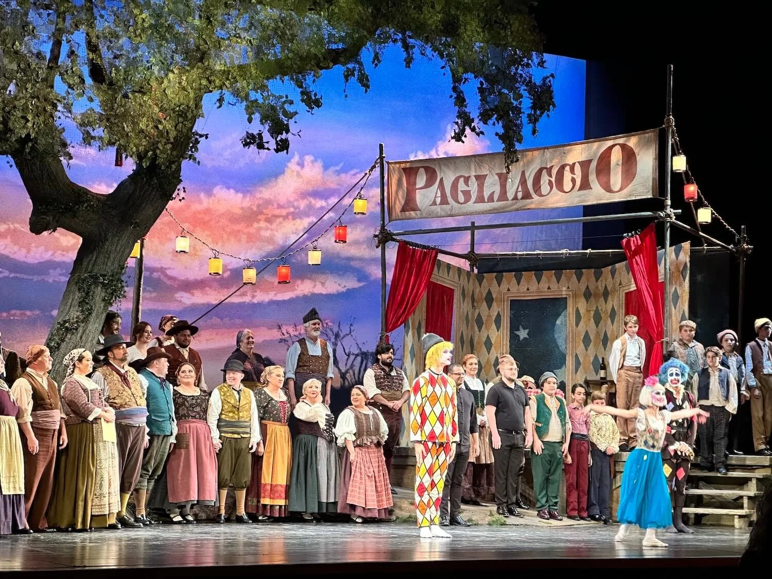 Cast of Pagliacci opera production on stage with large Pagliaccio banner, colorful costumes, string lights, and painted sky backdrop, featuring ballet dancer Emily Ricca in blue dress