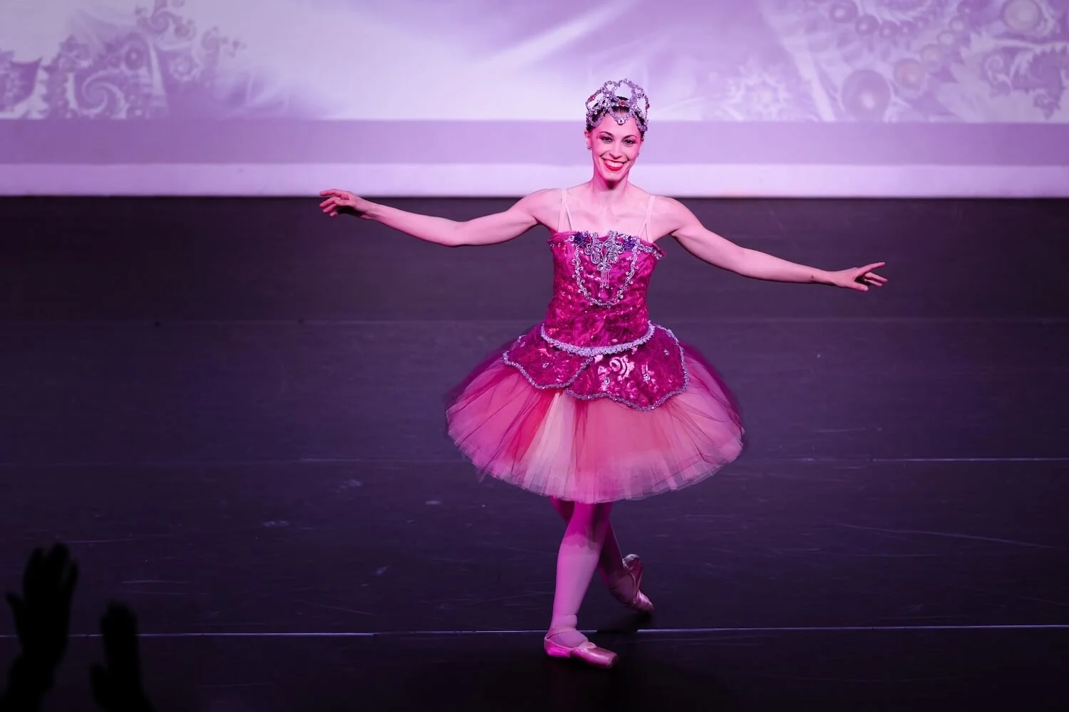 Ballet dancer Emily Ricca smiling on stage wearing a pink sequined tutu and crystal tiara during a solo performance under purple stage lighting