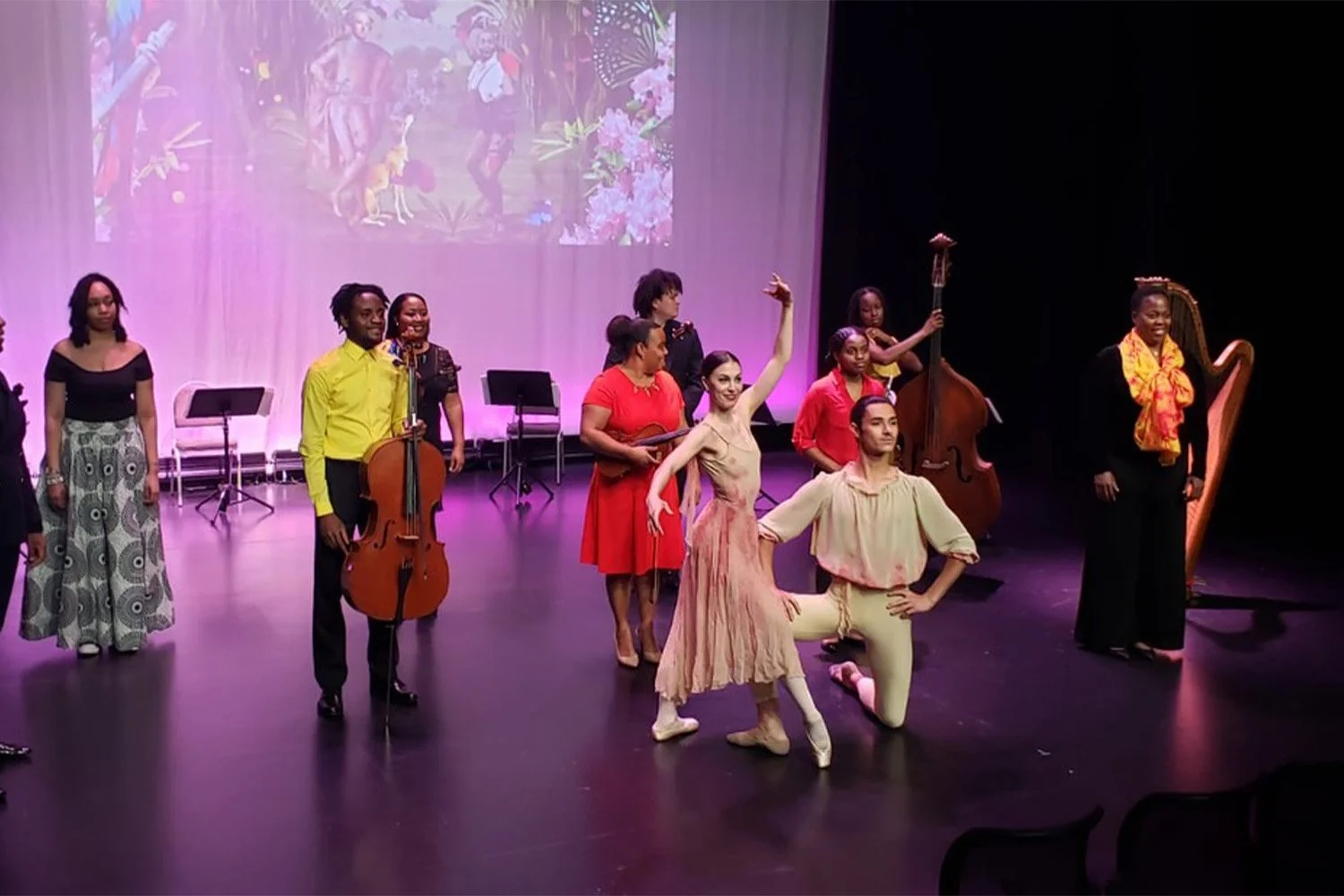 Ballet dancer Emily Ricca taking a bow on stage with male partner during curtain call, surrounded by live musicians including cellist, double bassist, and harpist under purple stage lighting with floral projection backdrop