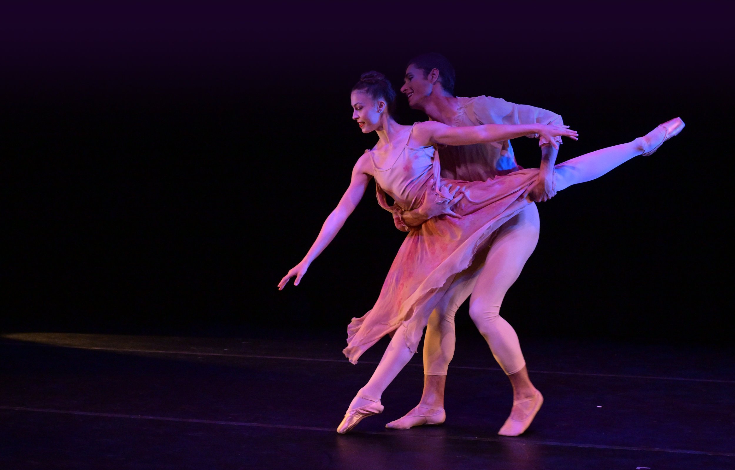 Two ballet dancers performing a dynamic pas de deux on stage, with the female dancer extending her leg in arabesque while being supported by her partner, both in soft pink costumes under warm stage lighting