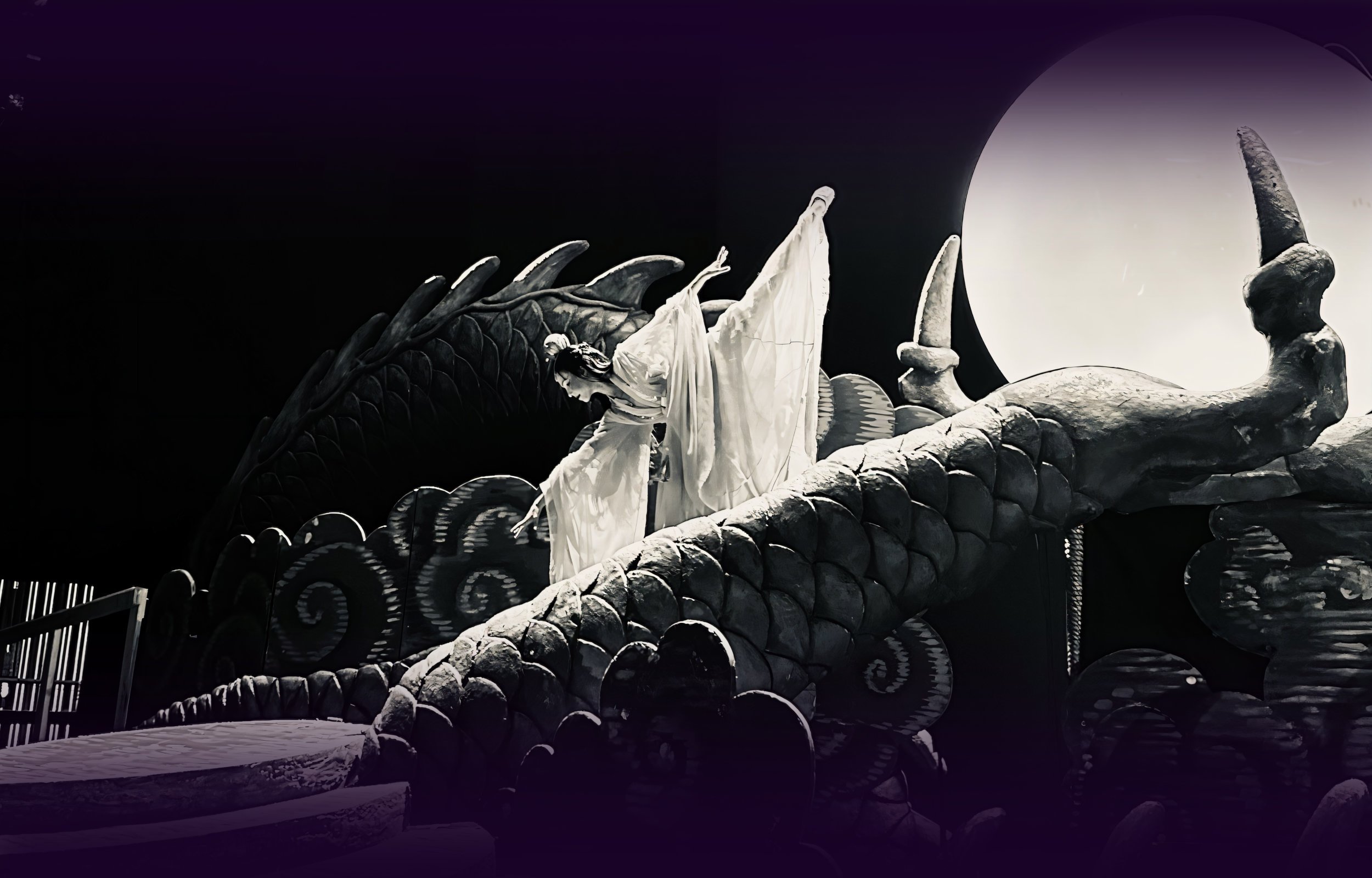 Ballet dancer Emily Ricca performing in Turandot opera, wearing a white flowing costume posed dramatically on a massive dragon stage prop beneath a glowing moon orb