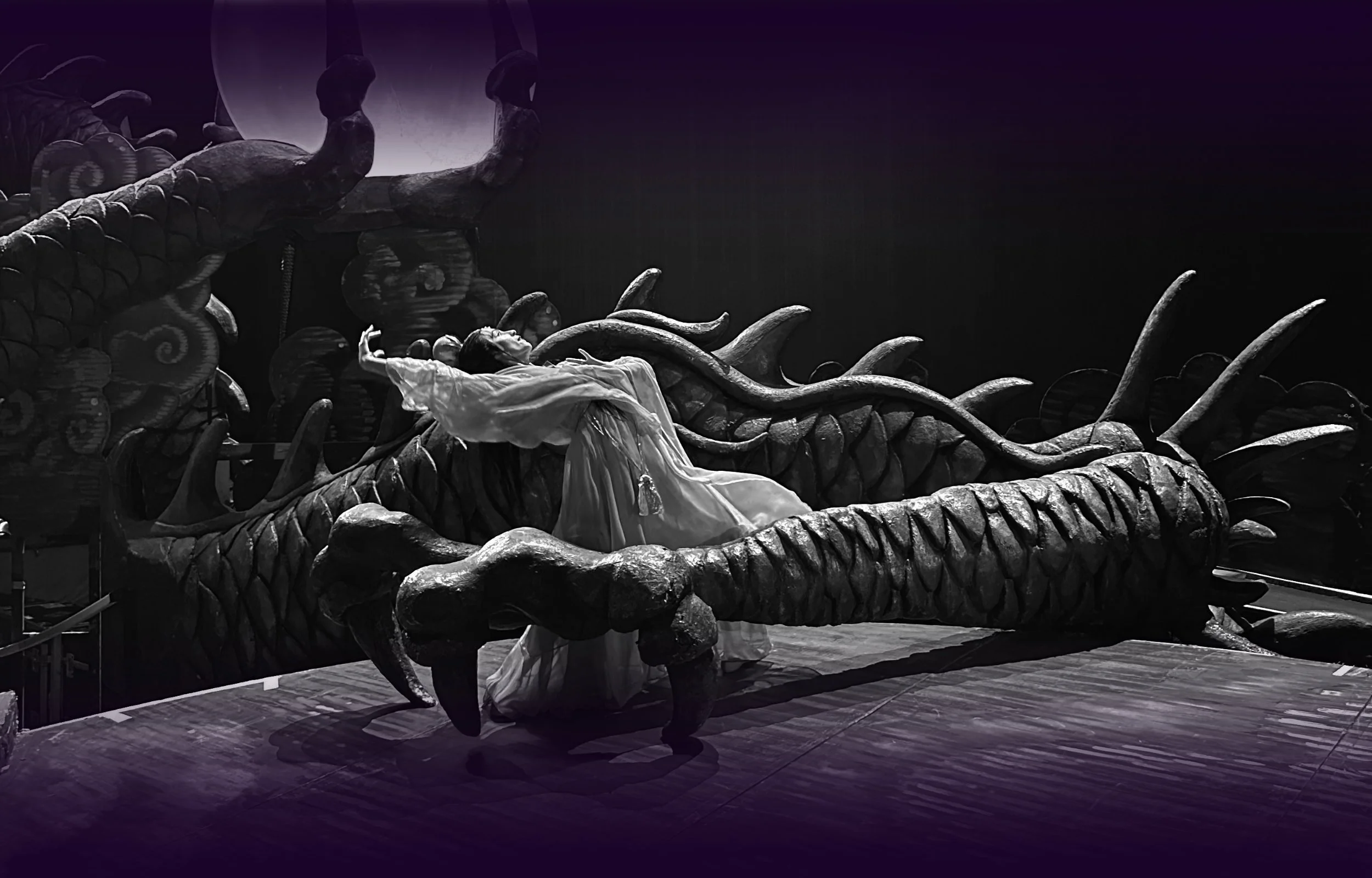 Ballet dancer in a white flowing costume performing an expressive dramatic pose beside a massive sculptural dragon set piece in a black and white stage production of Turandot