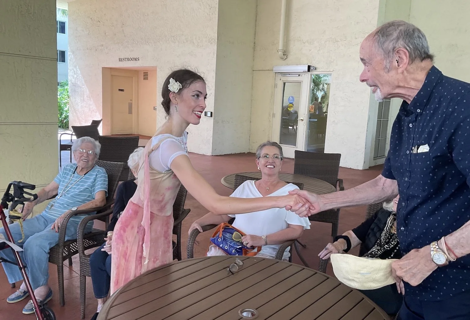 Ballet dancer Emily Ricca in costume warmly shakes hands with an elderly male resident at an outdoor senior living community gathering while others smile and watch
