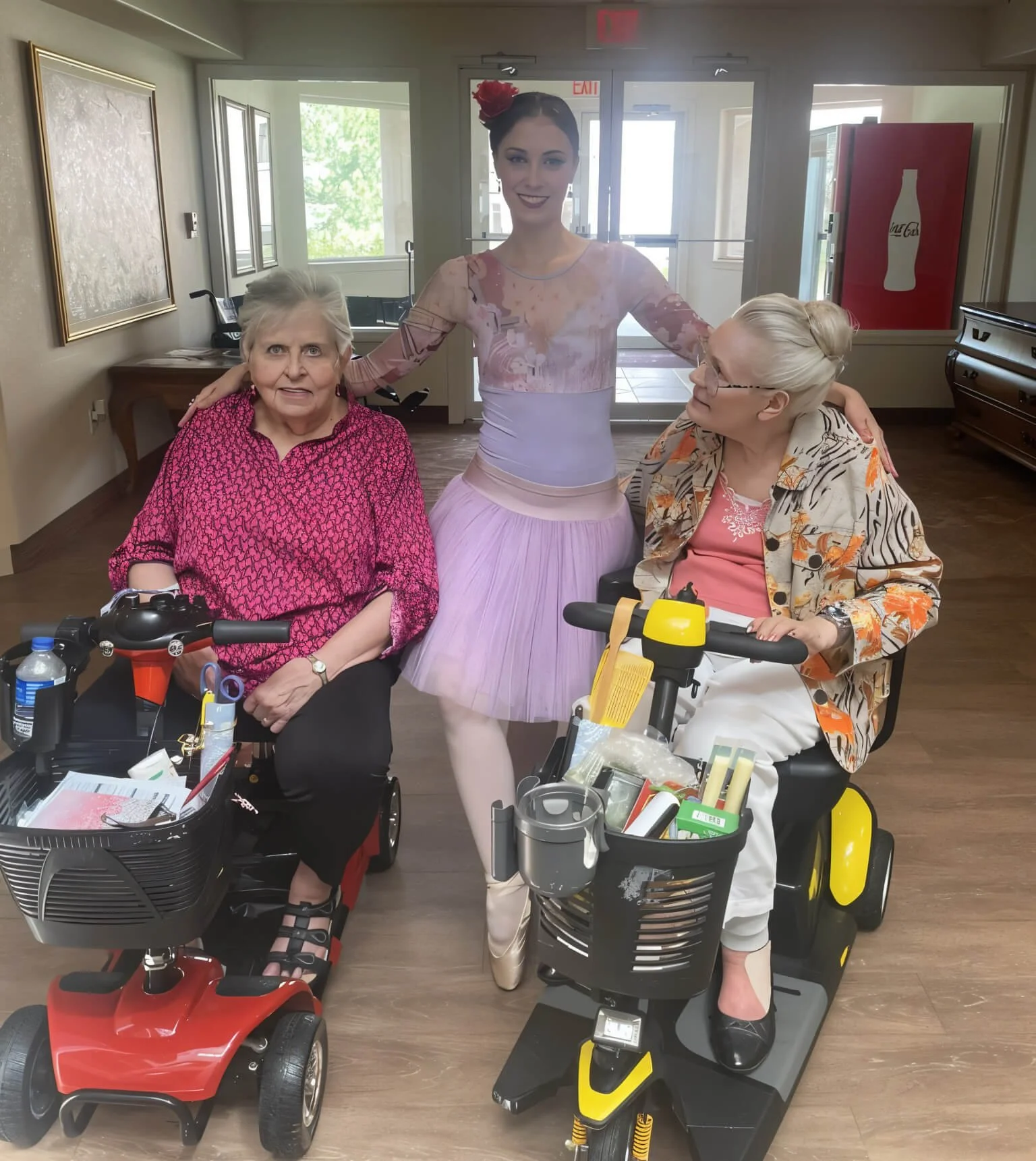 Ballet dancer Emily in pink tutu and pointe shoes poses with two elderly female residents on mobility scooters inside a senior living community common room