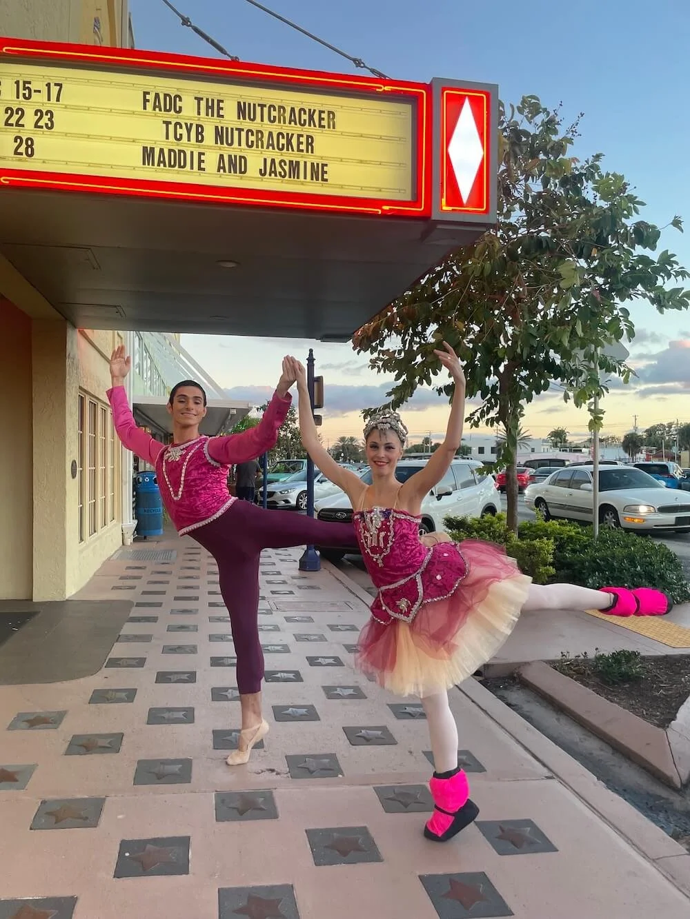 Ballet dancer Emily Ricca and male partner posing in pink and magenta Nutcracker costumes outside theater with FADC The Nutcracker marquee sign