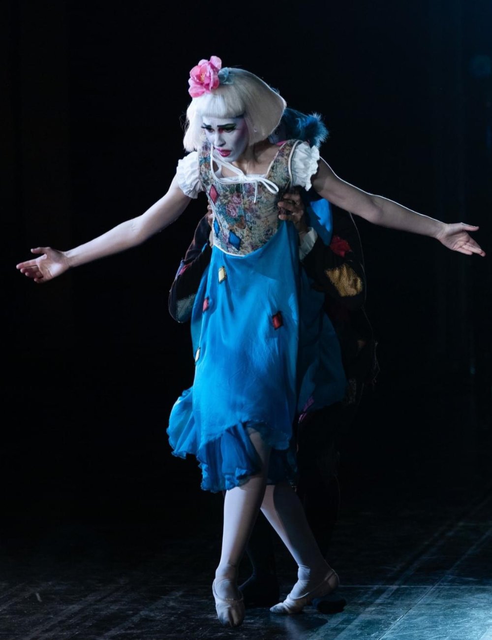 Ballet dancer Emily Ricca as Columbina in Pagliacci wearing a flowing blue dress, white theatrical makeup, and pink flower in hair on a dark dramatic stage