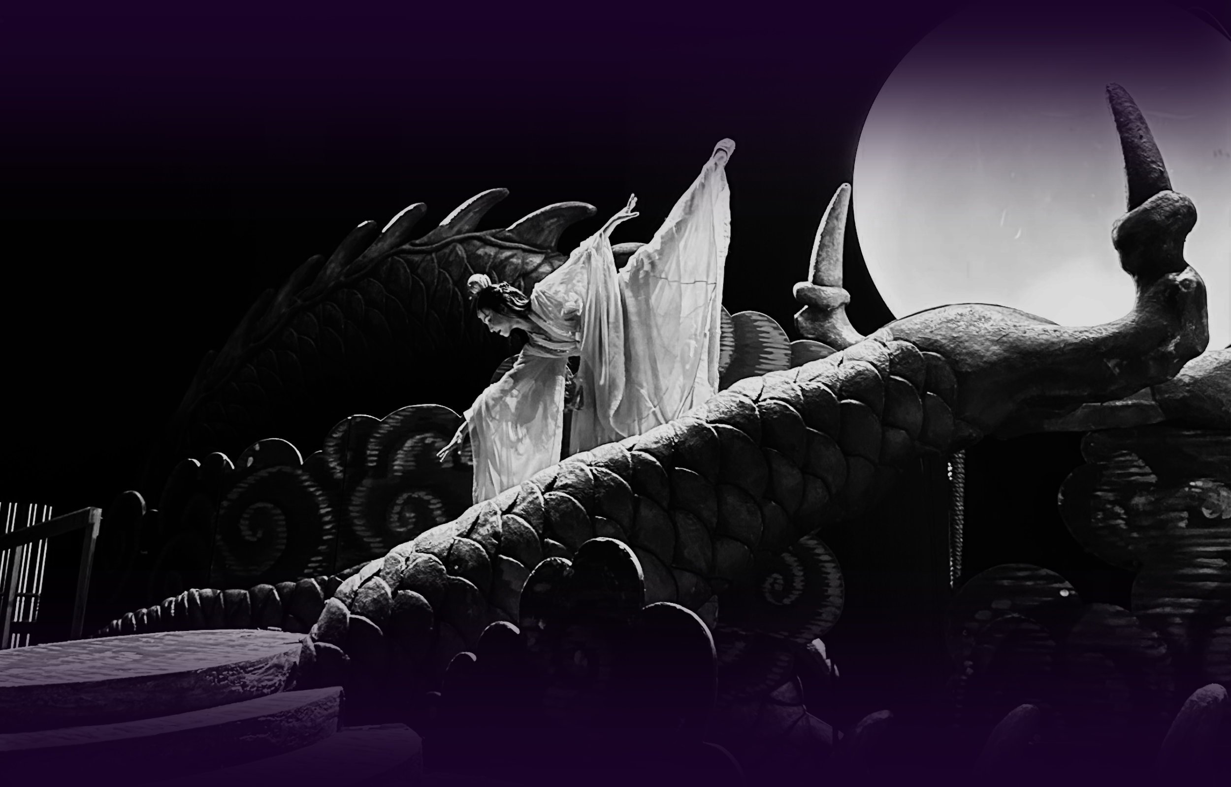 Ballet dancer Emily Ricca performing in Turandot opera, wearing a white flowing costume posed dramatically on a massive dragon stage prop beneath a glowing moon orb