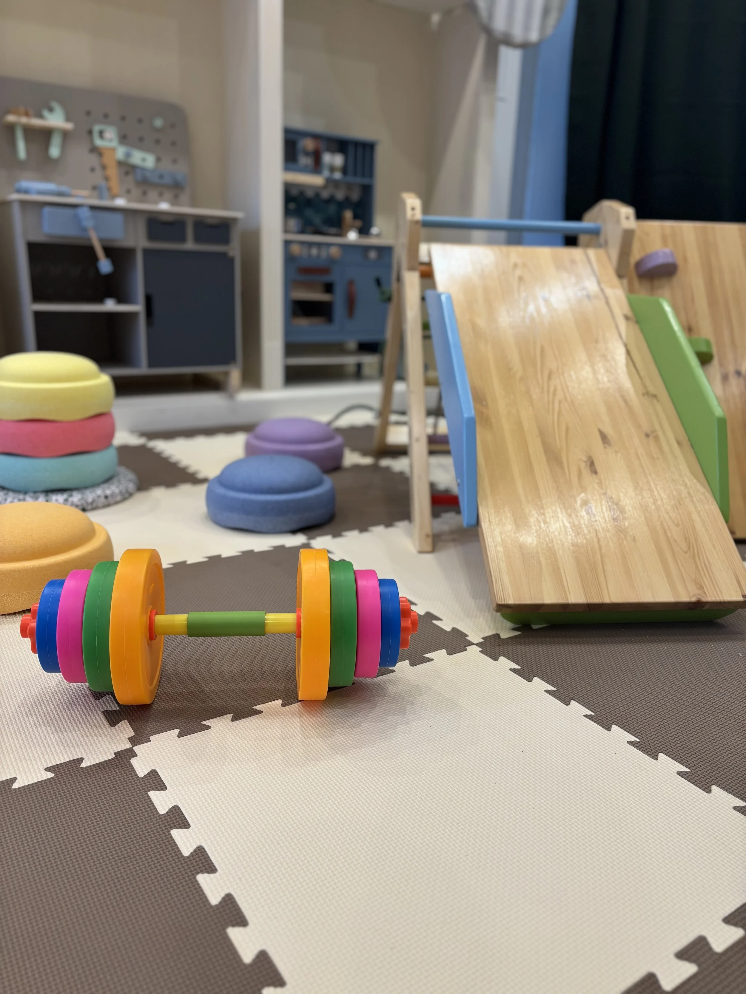 Kleiner Junge hebt mit beiden Händen eine Hantel im Spielzimmer. Im Hintergrund sind Kinder und Erwachsene, sowie bunte Bälle auf dem Boden.