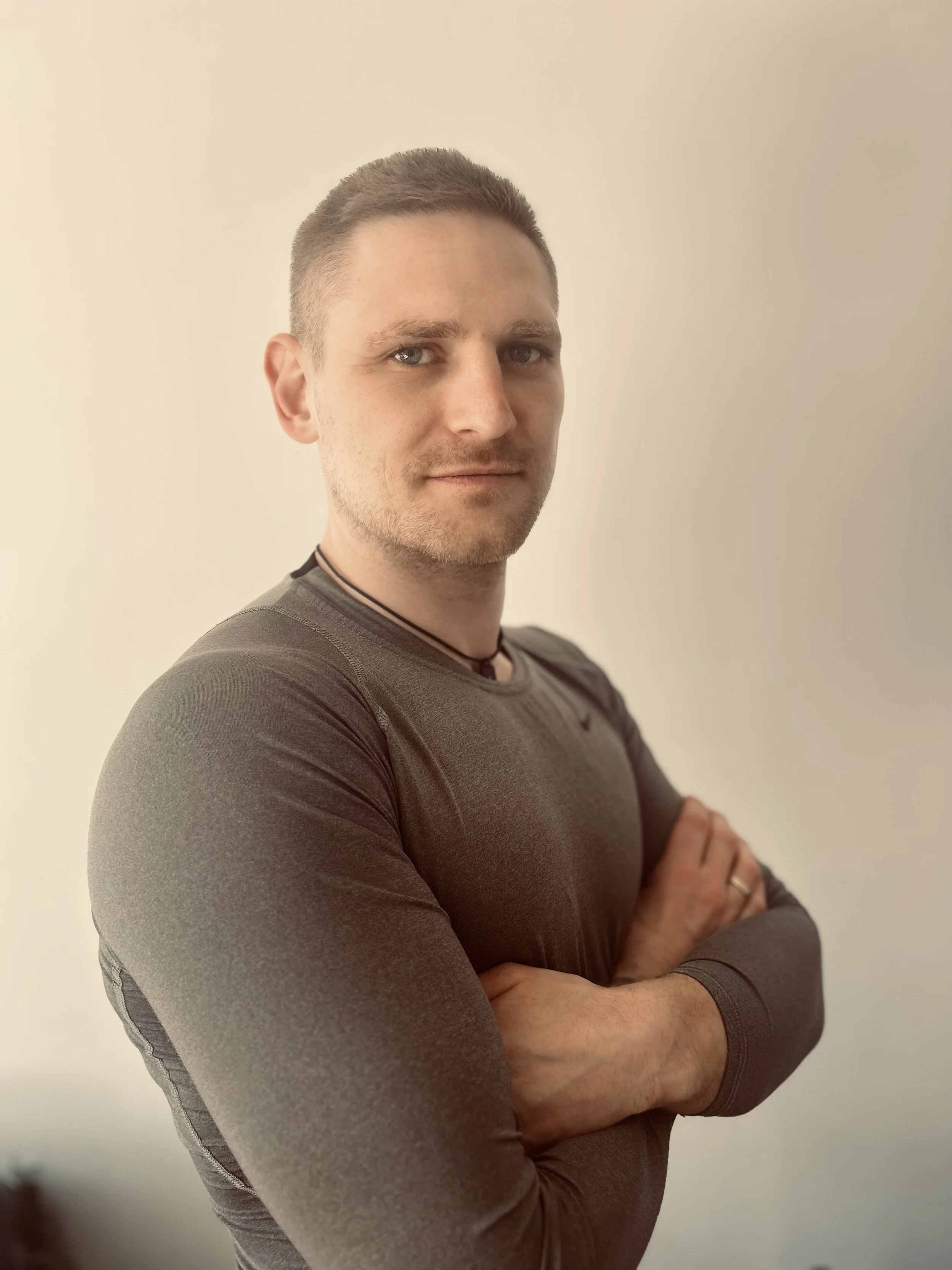 Porträt eines Mannes mit kurzen braunen Haaren, grauen Augen, graues Sportshirt, im Armkreuz, vor hellem Hintergrund.