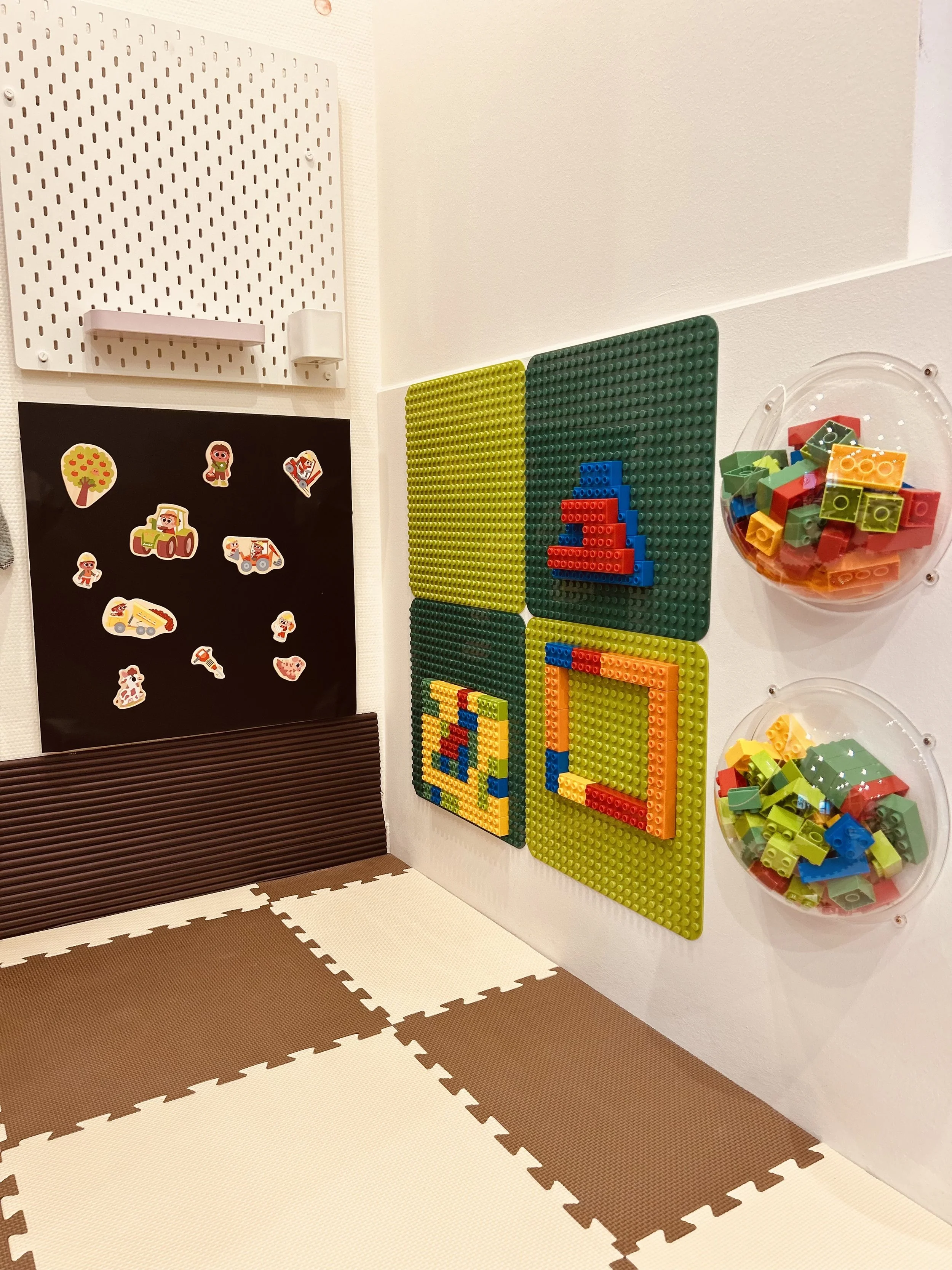 Spielzeugecke mit Lego und Aufklebern. Auf der Wand sind vier große Lego-Arrangements, darunter ein rotes und ein gelbes Quadrat, sowie eine Lego-Figur. Es gibt zwei klare Schalen mit Lego-Steinen und eine schwarze Tafel mit Aufklebern.