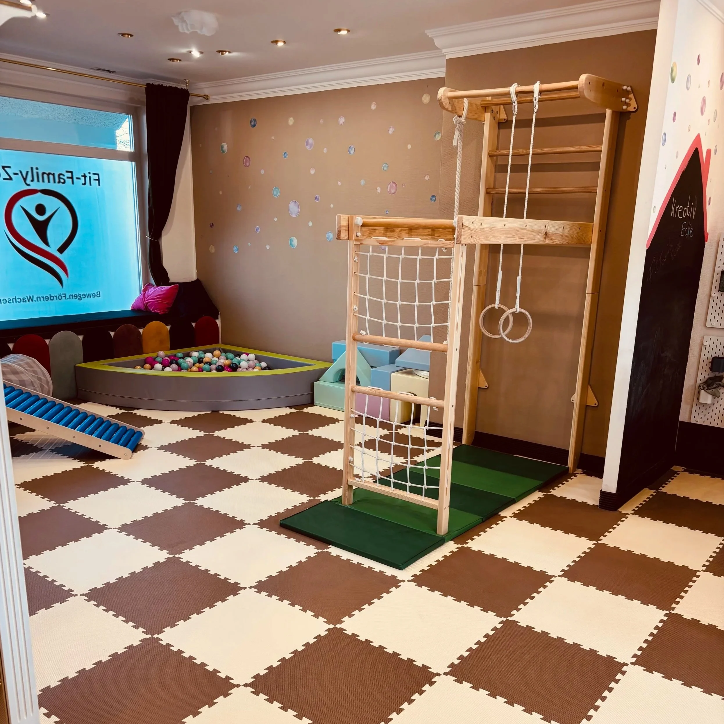 Ein Spielzimmer mit Klettergerüst, Rutsche, Bällebad und Wanddekoration. Es hat einen braunen Bodenbelag mit Schachbrettmuster und eine große Fensterfront.