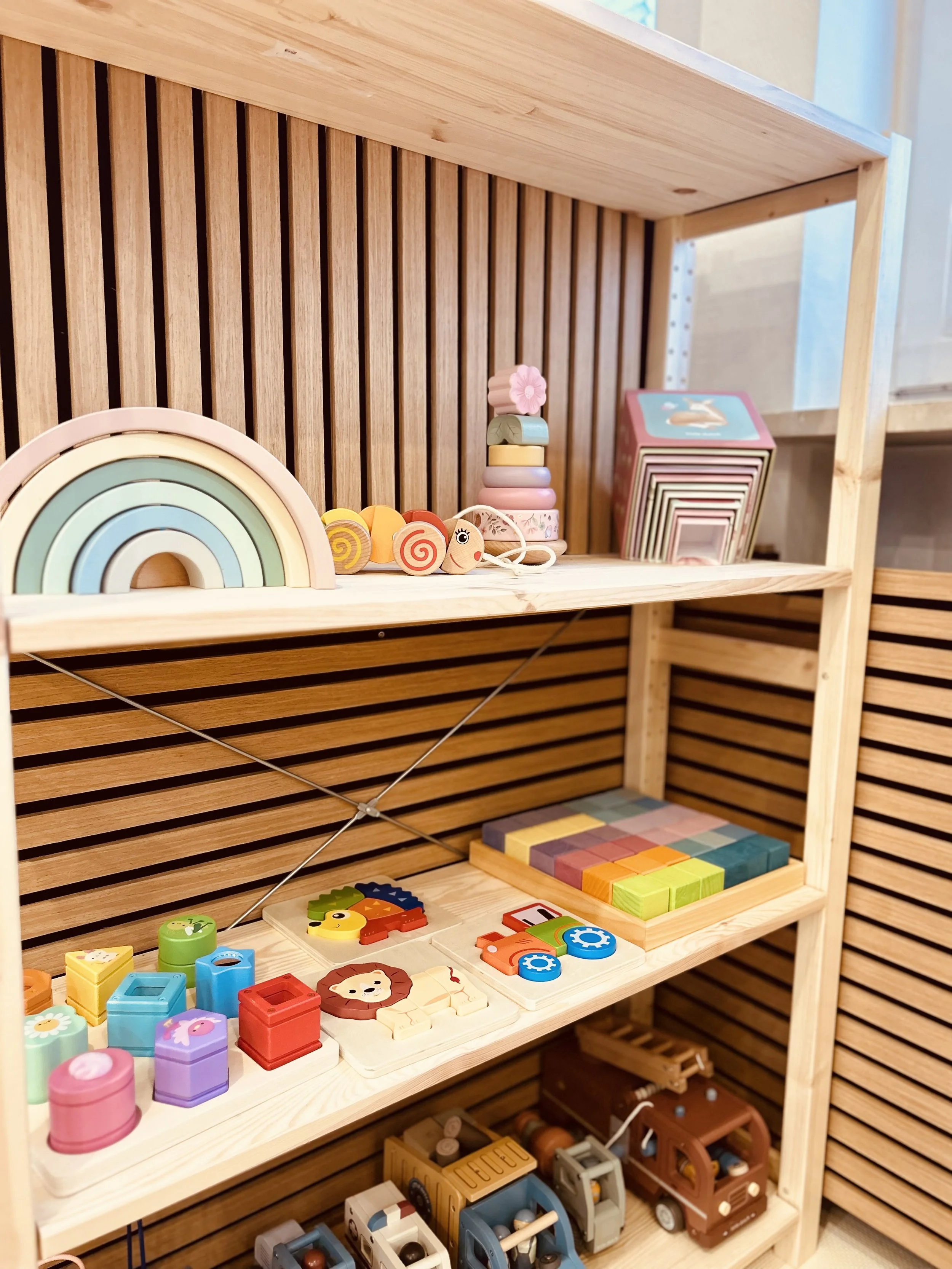 Holzregale mit buntem Holzspielzeug, einschließlich Regenbogen, Stapelringe, Tierknochen, Würfel, Autos und andere Figuren.