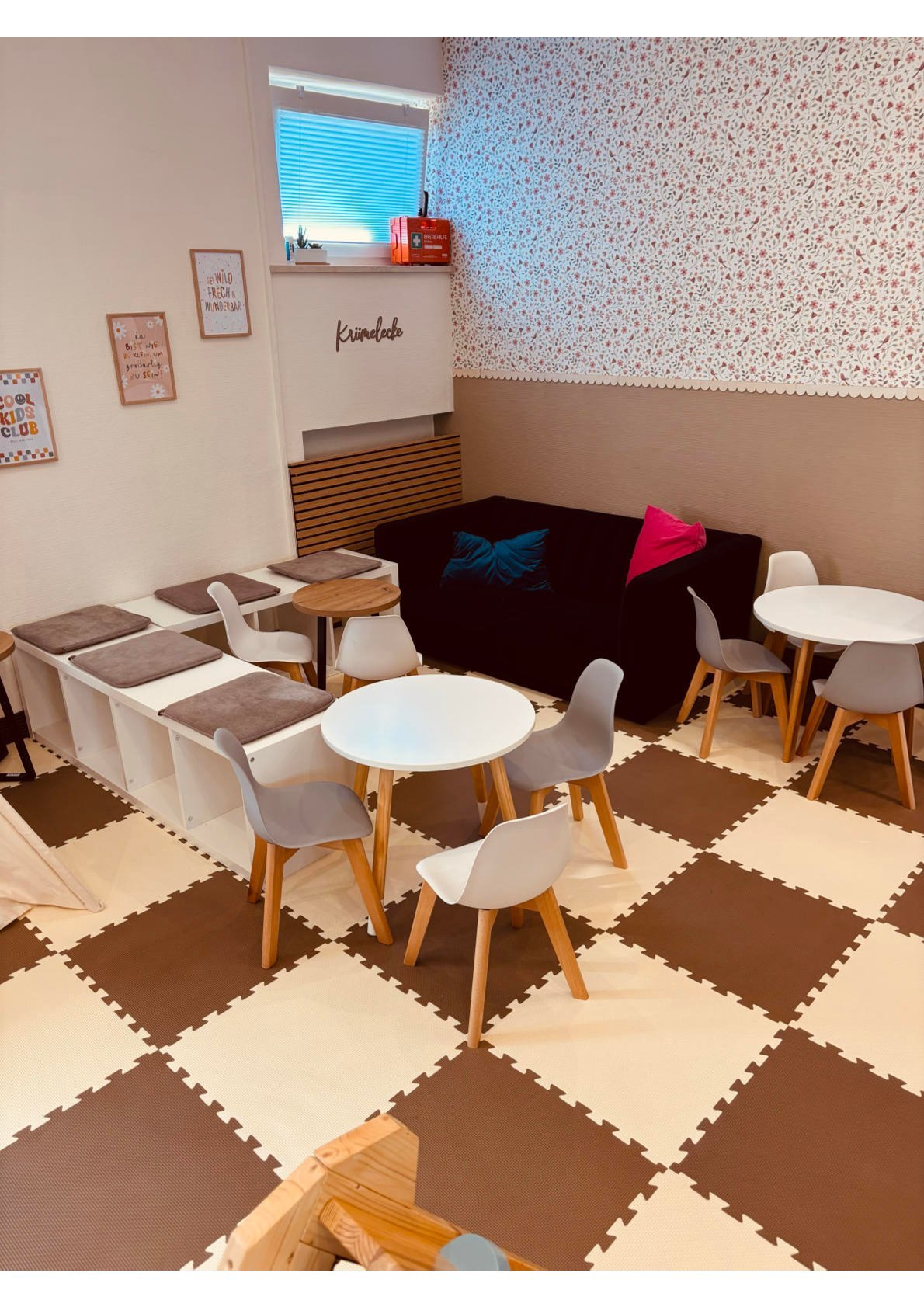 Ein Kinderwartezimmer mit Tischen, Stühlen, einem schwarzen Sofa mit bunten Kissen, buntem Pflasterboden, Wanddekoration und einem Fenster.