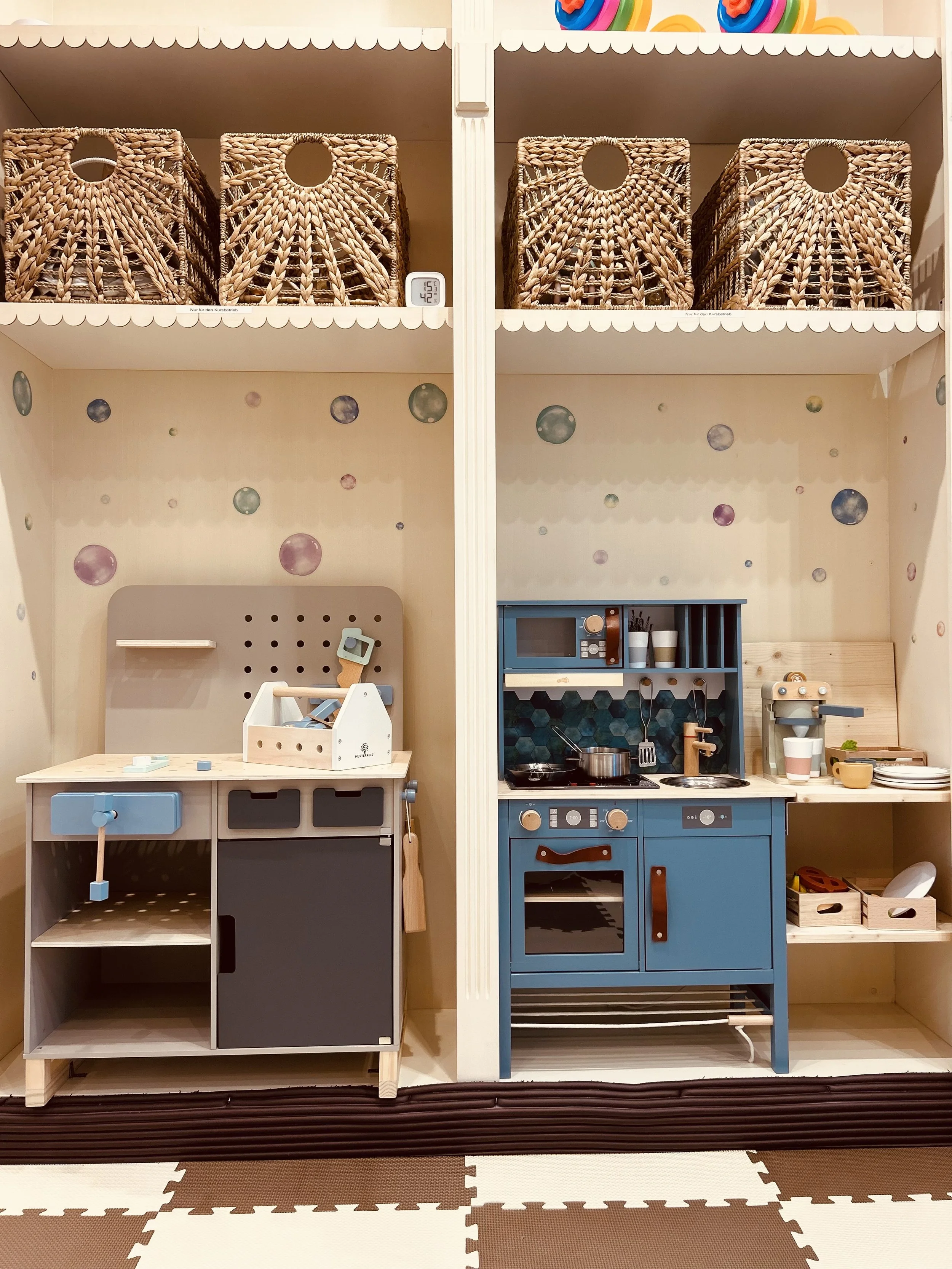 Miniaturküche mit blauer und grauer Ausstattung, Spielzeugkochtöpfe, Geschirr und Zubehör, im Hintergrund bunte Wanddekoration mit bunten Kugeln, zusammen mit einem Puppenhaus auf Regal über der Küche.