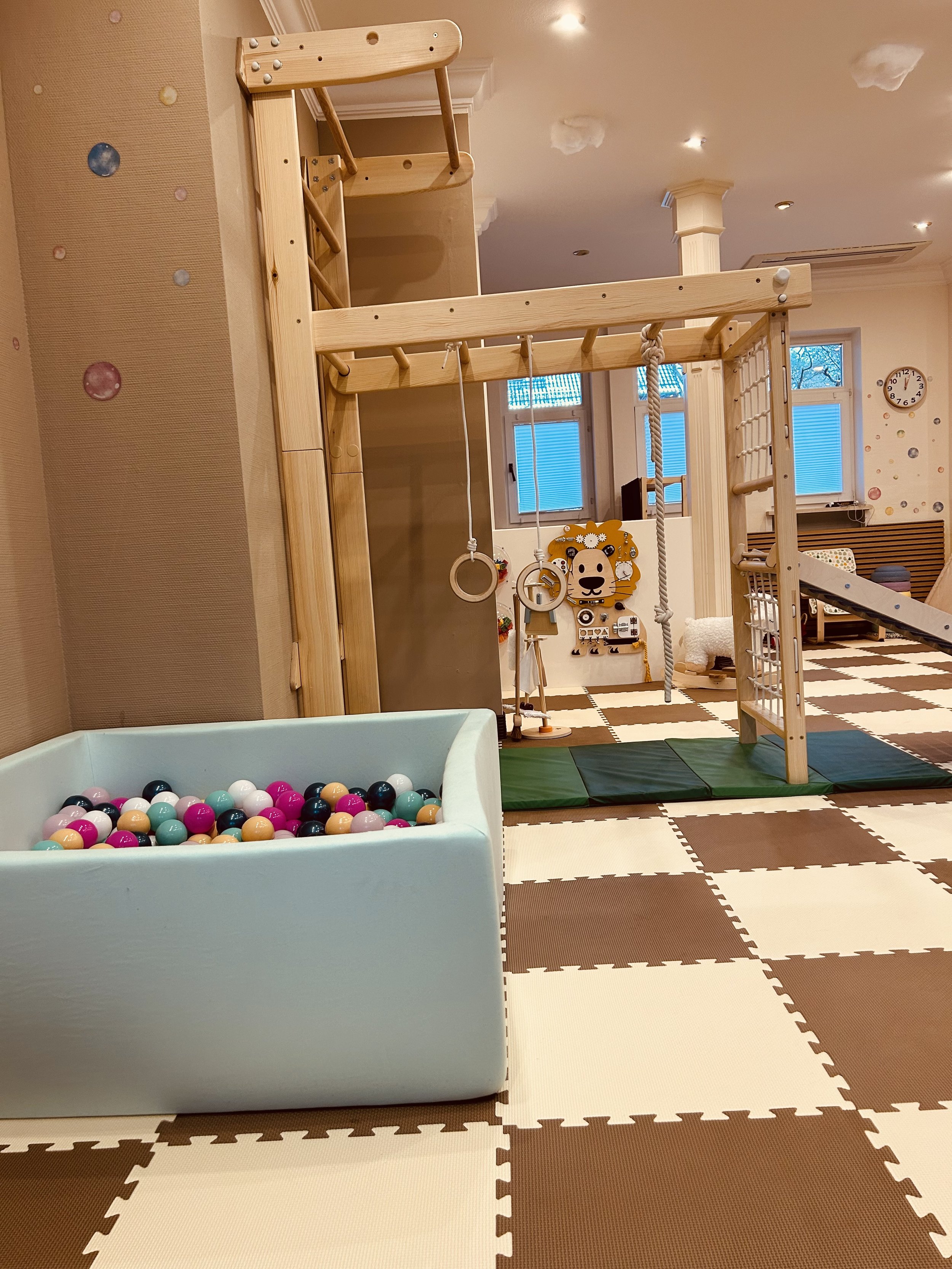 Innenraum eines Spielzimmers mit Klettergerüst, Ringen, Seil und Matten, ein Ballbad mit bunten Bällen, und eine Wanddeko mit Tiergesicht und Sonnenbrille.