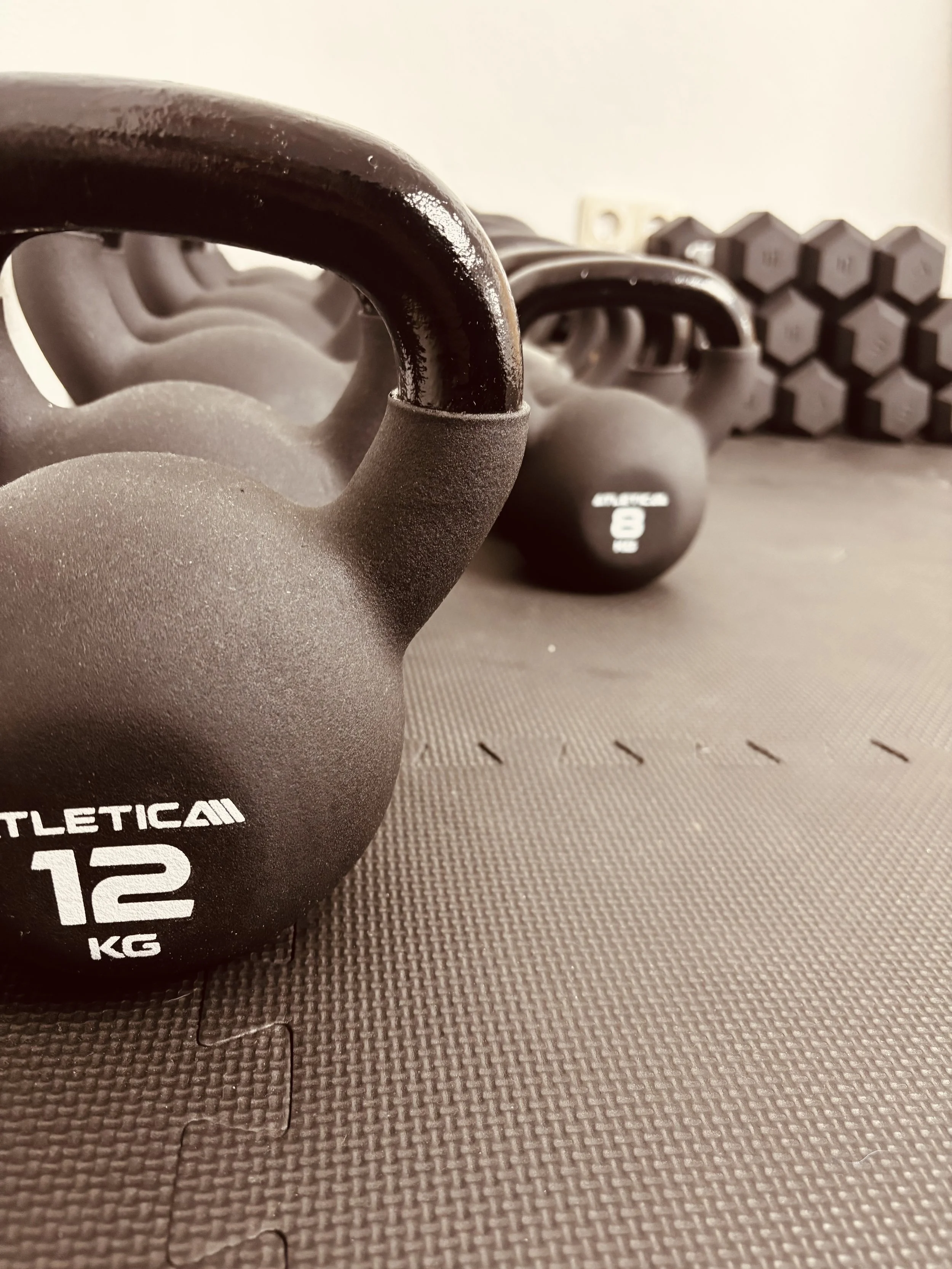 Mehrere schwarze Kettlebells mit 12 kg Gewicht auf einer Gymnastikmatte, im Hintergrund Hantelscheiben.