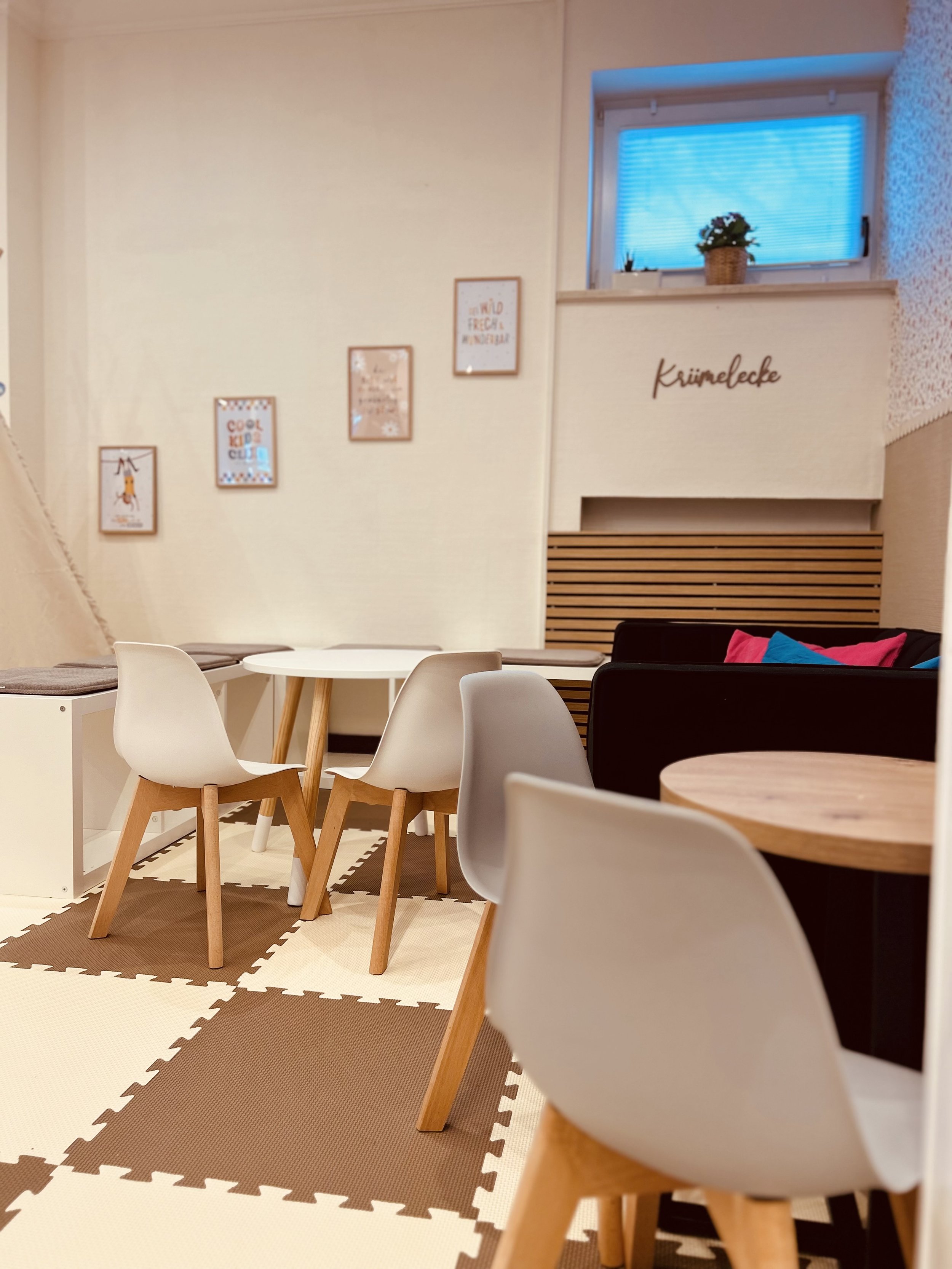 Ein gemütliches Kinder- und Familiencafé mit weißen Stühlen, kleinen Tischen, gemusterter Teppichboden in Braun und beige, dekorativen Bildern an der Wand und einem blauen Licht durch das Fenster. Es gibt eine Plattform mit dem Schriftzug 'Krämelcke'