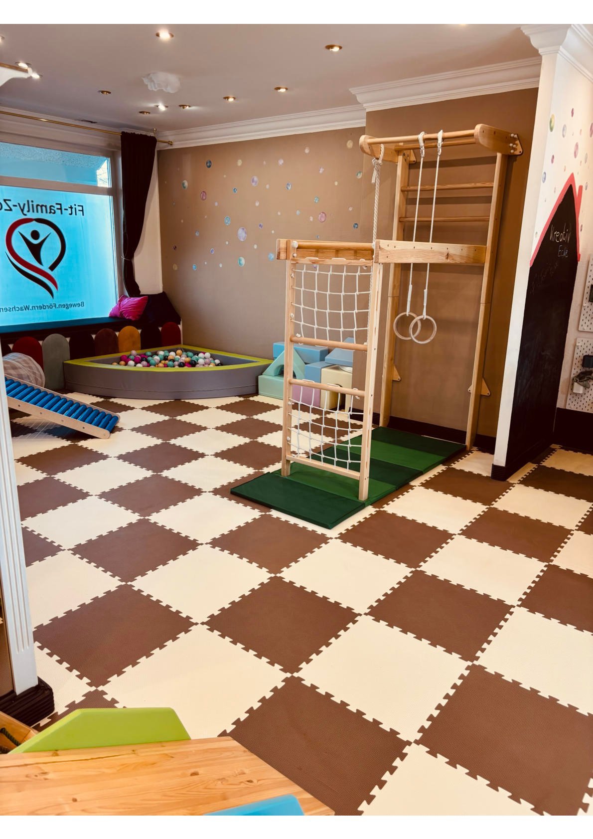 Spielplatz in einem Spielzimmer mit Klettergerüst, Ballgrube, Spielelementen und schwarzen und weißen Teppichfliesen.