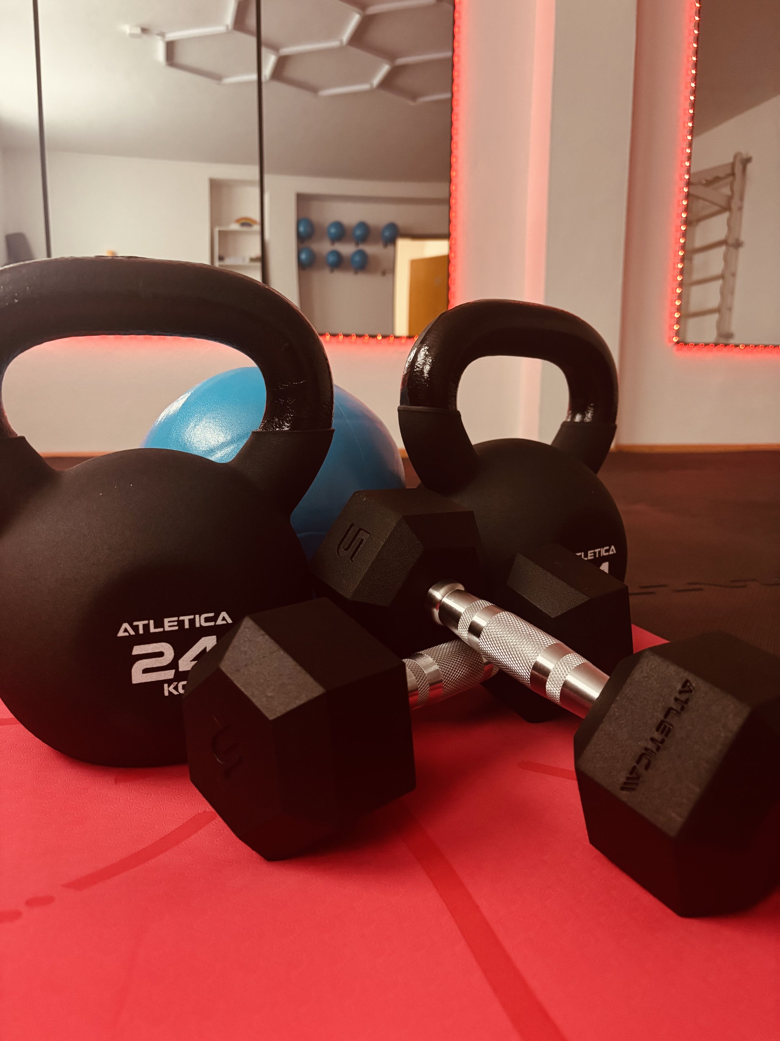 Fitnessgeräte im Fitnessraum, darunter zwei schwarze Kettlebells, zwei schwarze Kurzhanteln, eine blaue Gymnastikball, ein Spiegel mit roter LED-Beleuchtung, eine Gymnastikmatte auf dem Boden und weitere Fitnessgeräte im Hintergrund.
