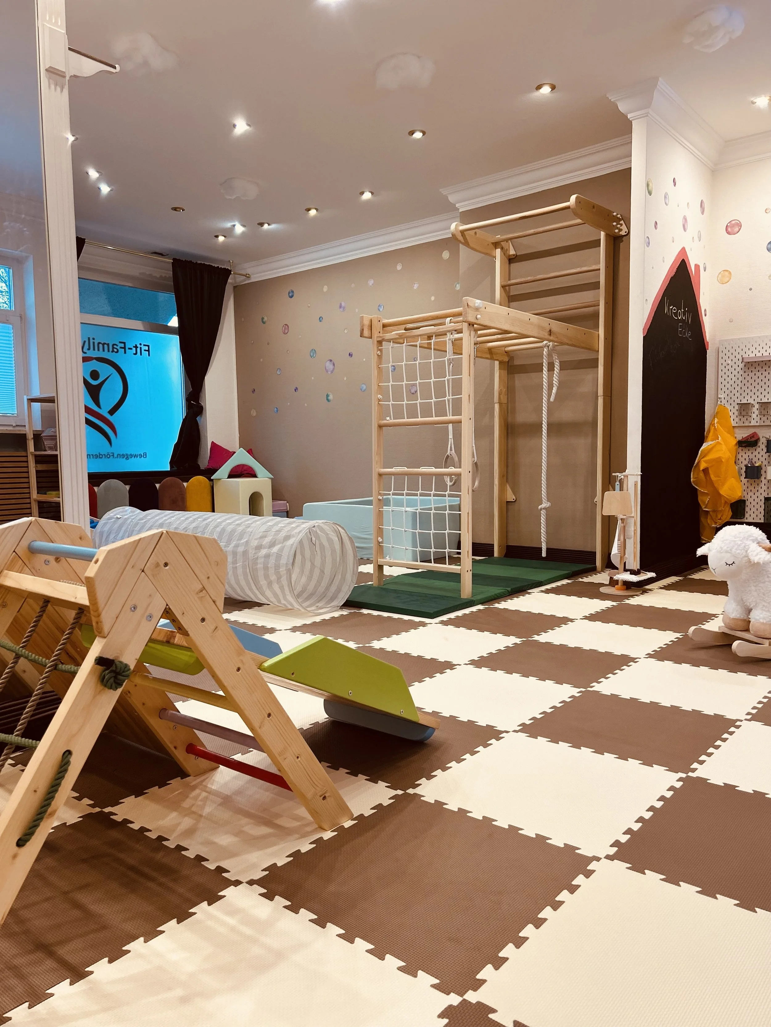 Kleiner Junge hebt mit beiden Händen eine Hantel im Spielzimmer. Im Hintergrund sind Kinder und Erwachsene, sowie bunte Bälle auf dem Boden.