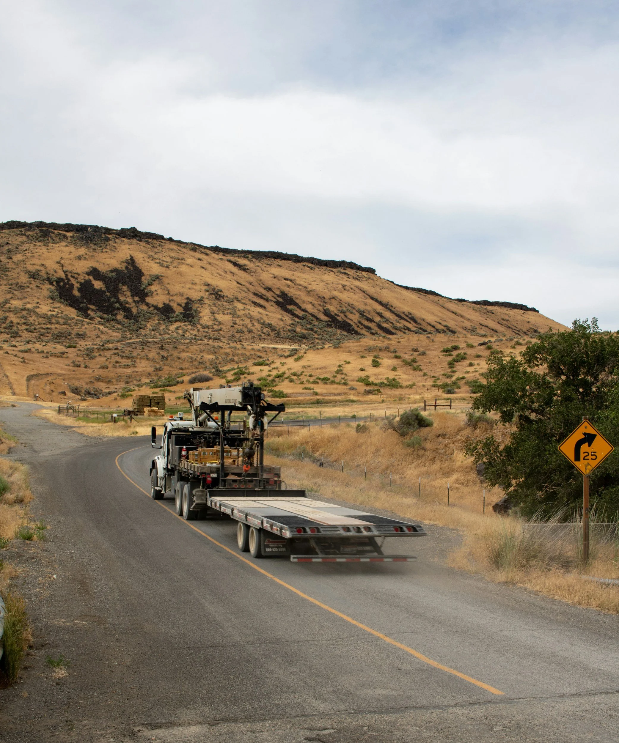 A large flatbed truck driving on a winding road through a rural, hilly landscape with dry grass, a tree on the right, and a yellow road sign indicating a sharp curve ahead.