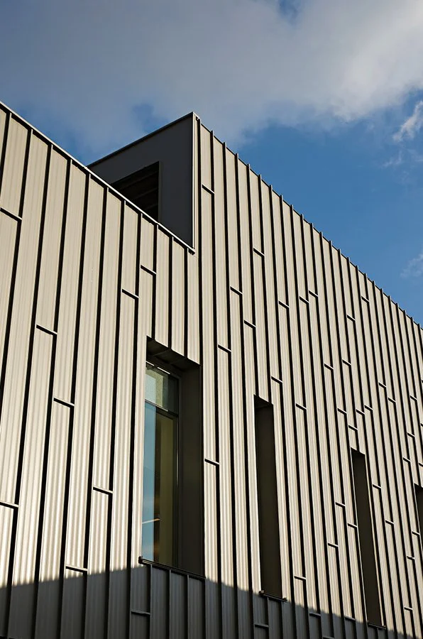 Close-up of a modern building's exterior with vertical metal panels and tall narrow windows against a partly cloudy sky.