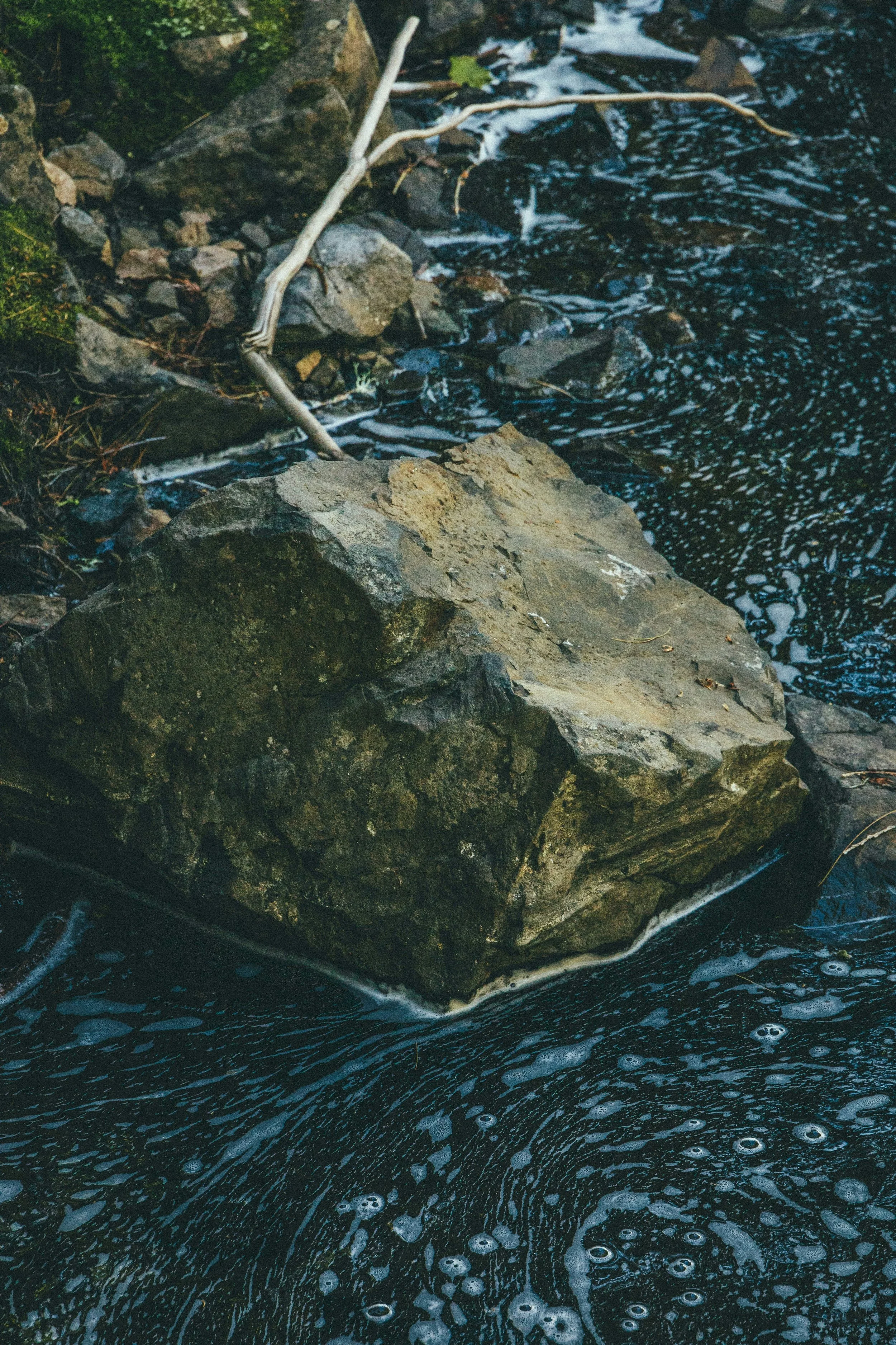 Un gros rocher au bord d'une rivière avec des galets et un bâton blanc posé dessus, entouré d'eau noire avec des bulles.