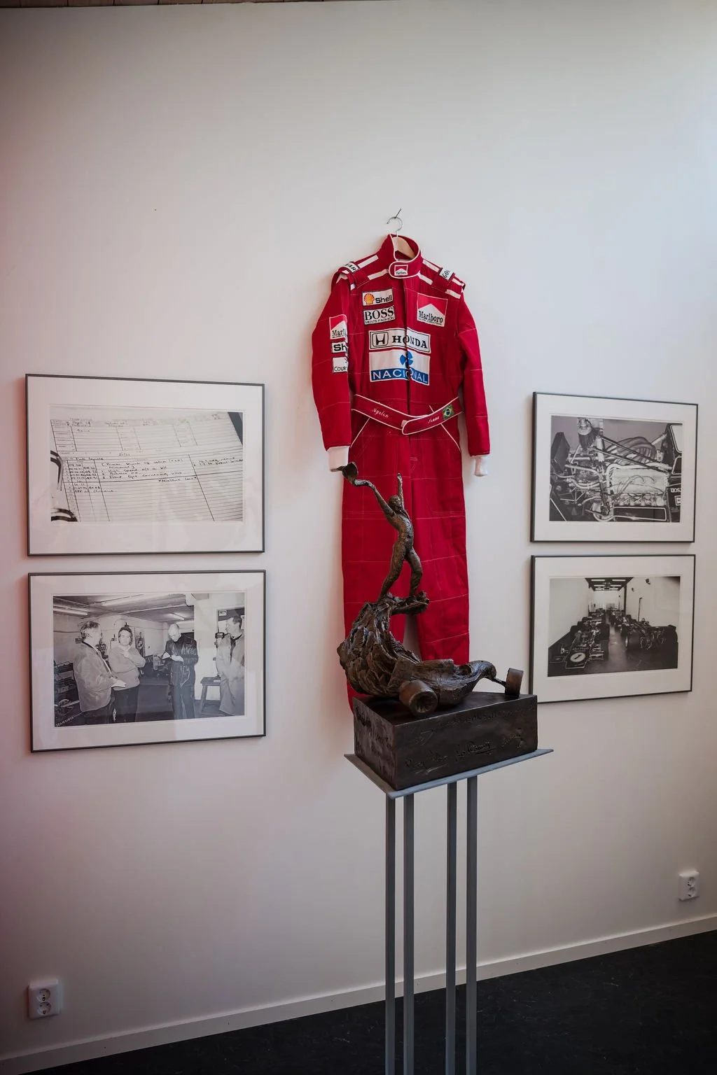 A red racing suit with sponsor patches hanging on a white wall, a bronze sculpture of a man in a dynamic pose on a pedestal, and four black-and-white framed photographs related to racing and racing teams.