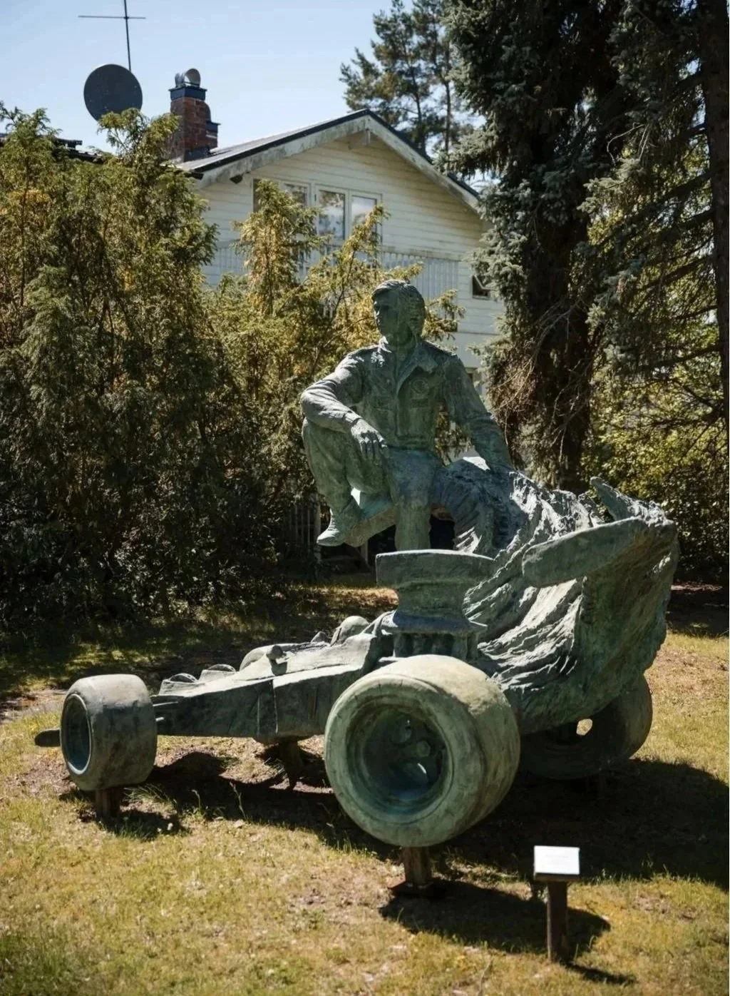 Bronze statue of a person sitting on a disassembled go-kart, outdoors with trees and a house in the background.