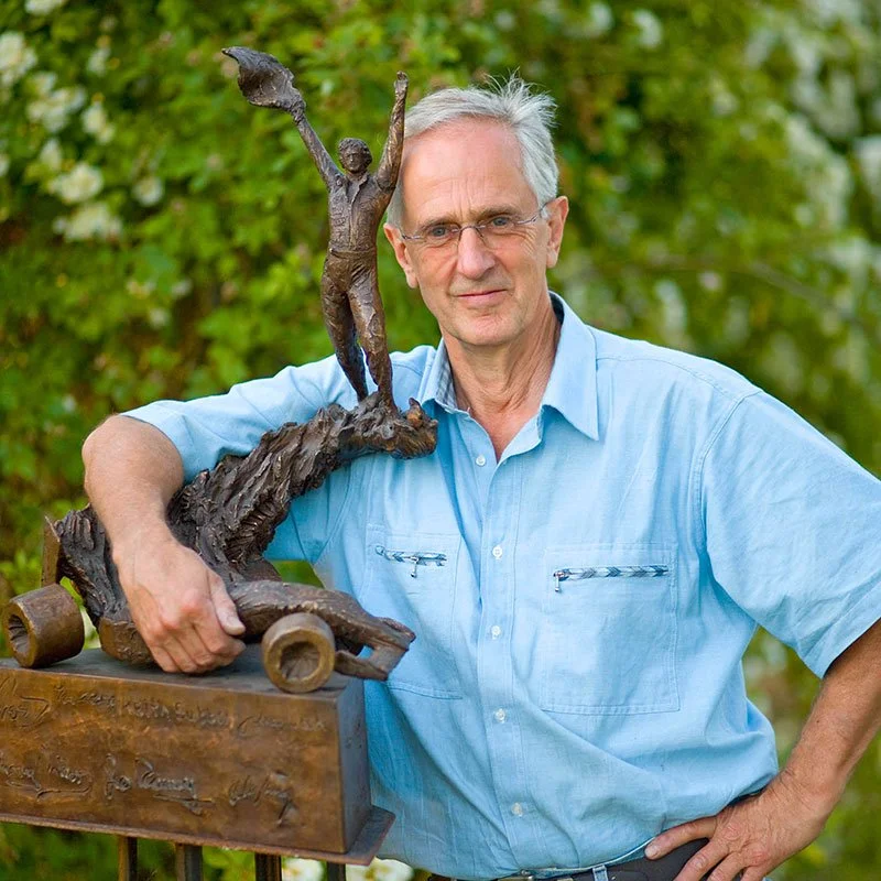 An older man in a light blue shirt standing outdoors with greenery in the background, holding a bronze sculpture of a person with arms raised.